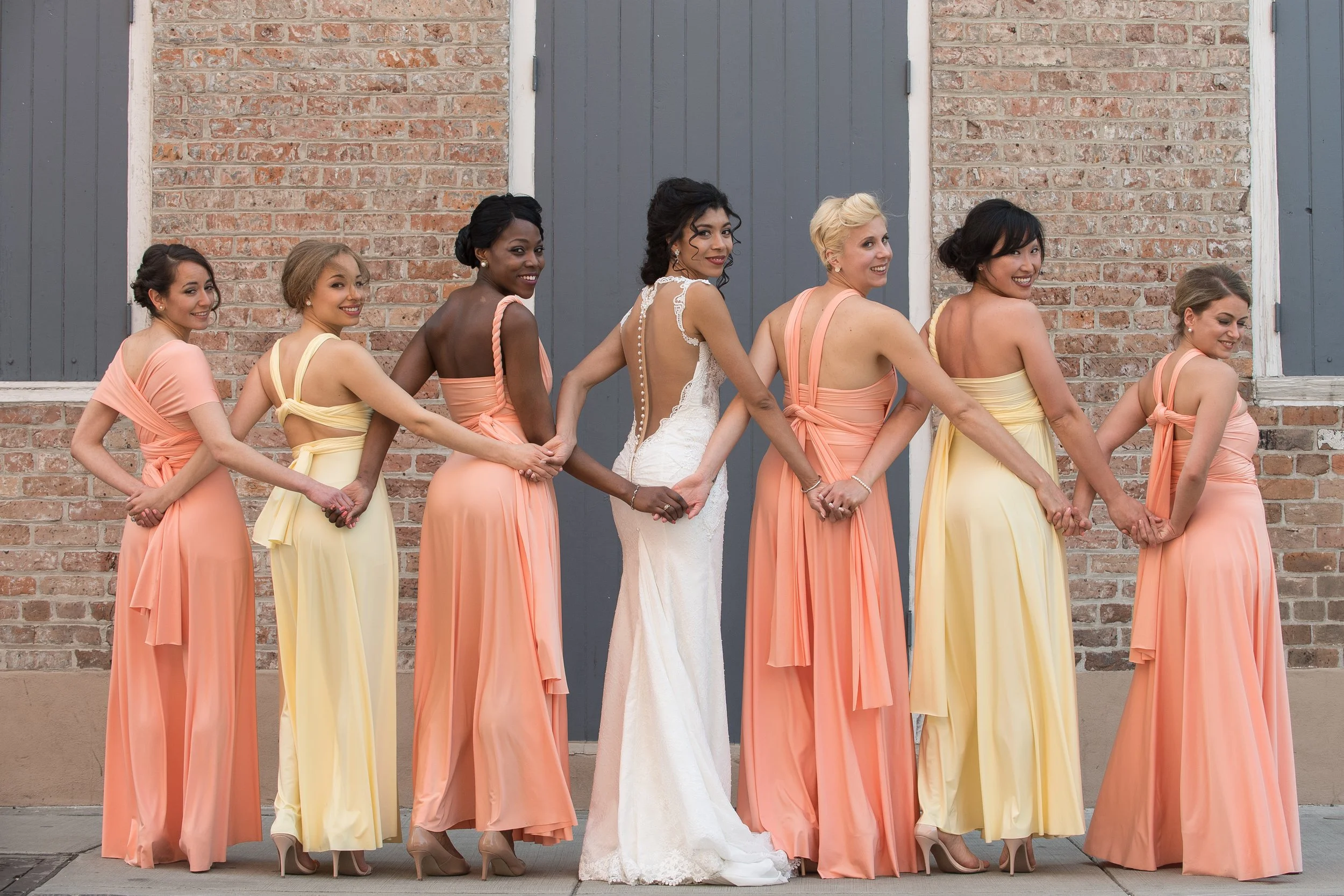 A diverse group of eight women dressed in pastel-colored dresses holding hands in front of a brick wall with gray doors. One woman in the center is wearing a white wedding gown, indicating a wedding celebration.