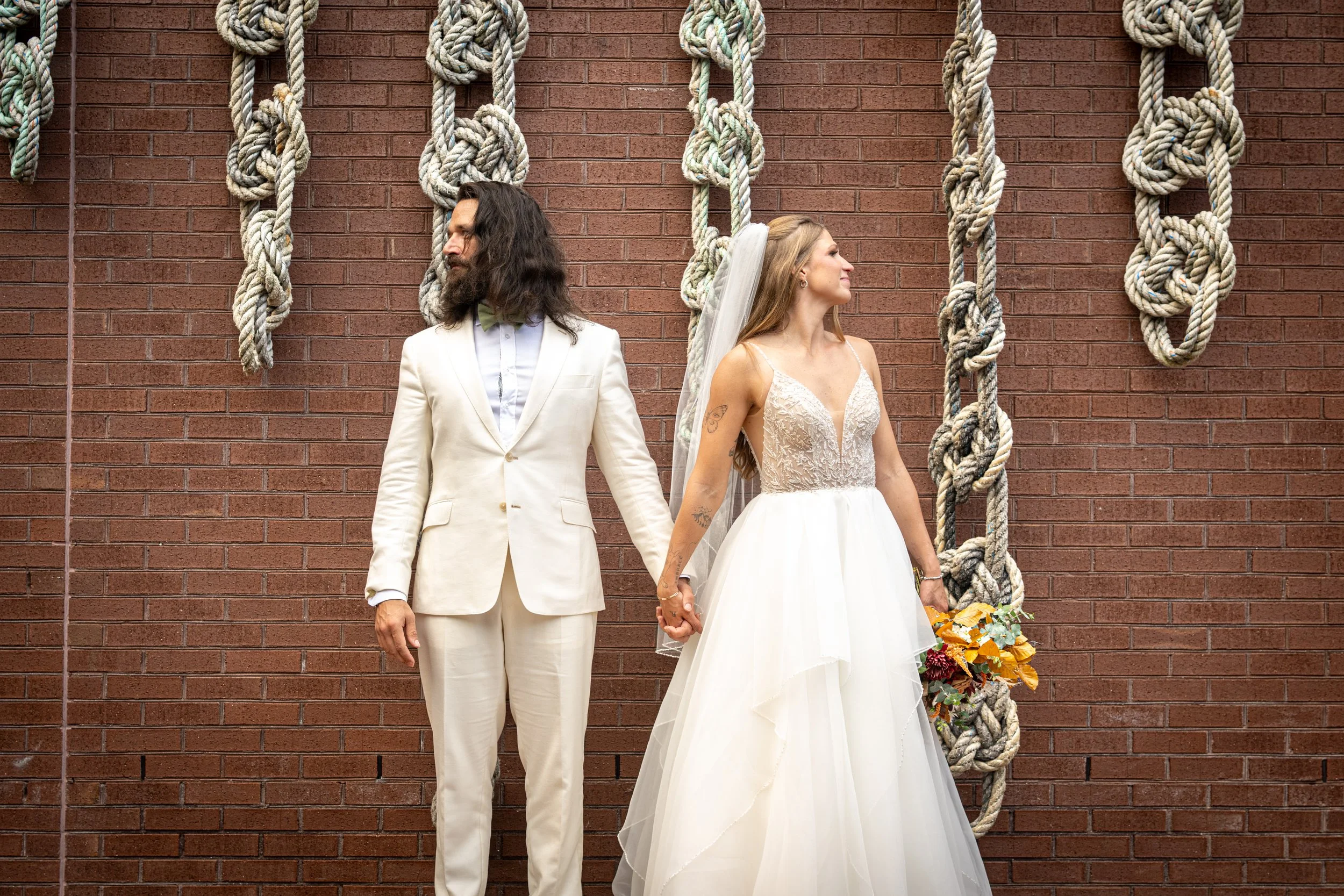 A couple dressed in wedding attire stands hand in hand against a brick wall decorated with thick ropes forming decorative knots.