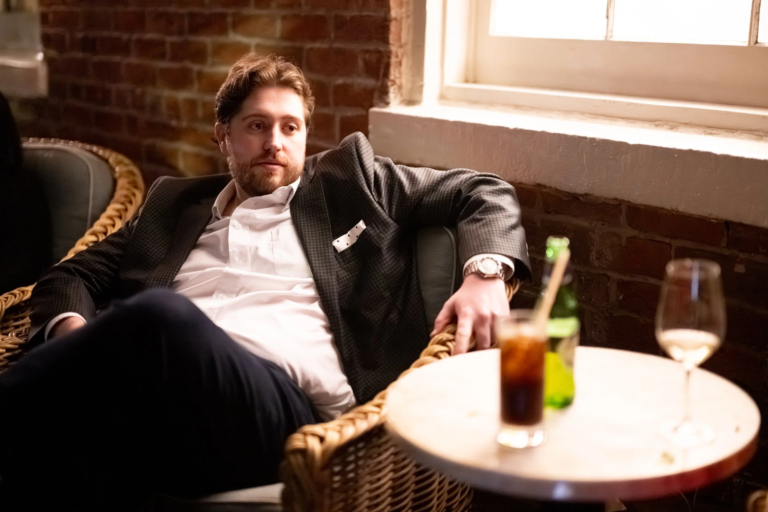 A man with brown hair and a beard is sitting on a wicker chair in a restaurant or bar with brick walls and a large window. He is dressed in a dark plaid suit with a white shirt and a pocket square, and is wearing a watch. On the table in front of him