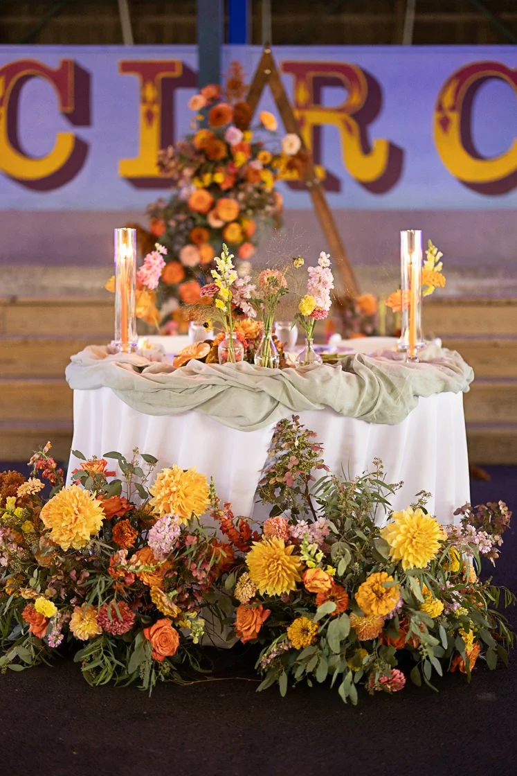Decorative table with floral arrangements and candles in glass holders, set up for a celebration or event, with a colorful banner and flower arrangements in the background.