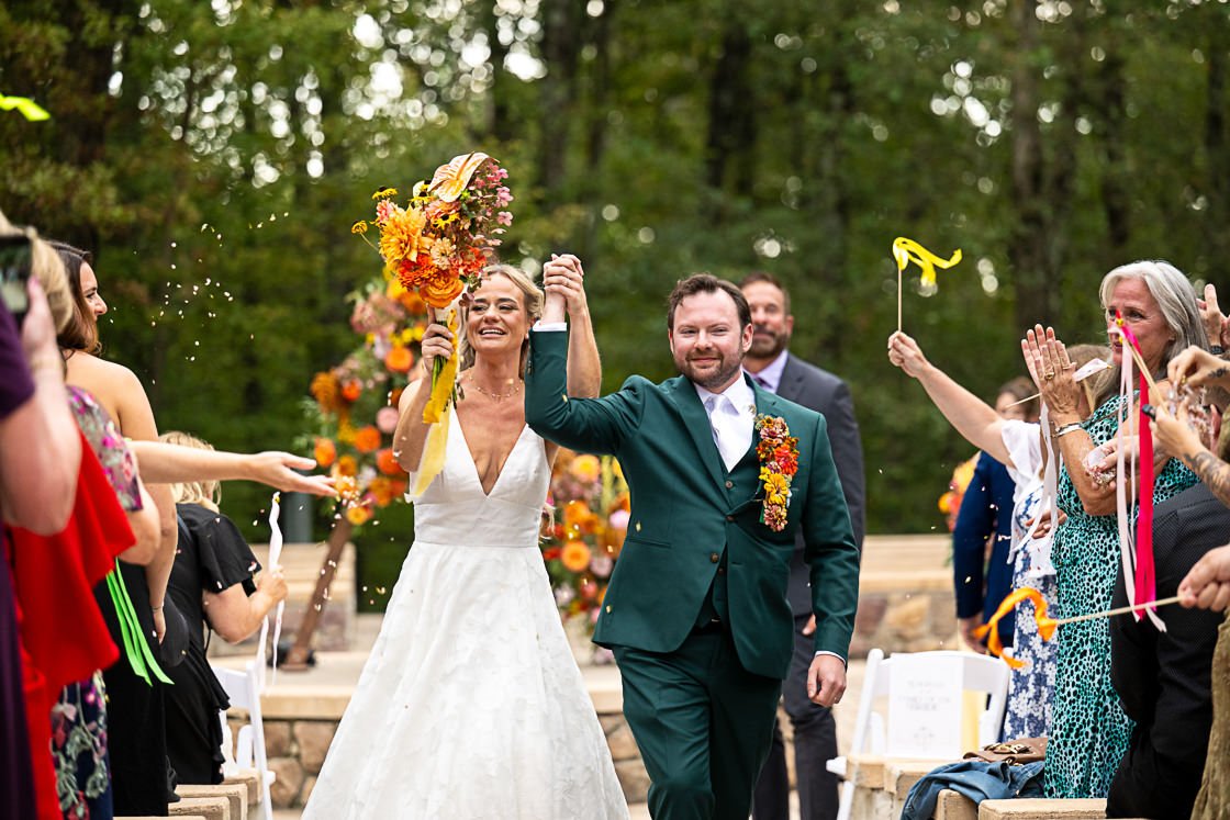 A newlywed couple celebrating their wedding outdoors, walking hand in hand, surrounded by friends and family, with a flower arch in the background.
