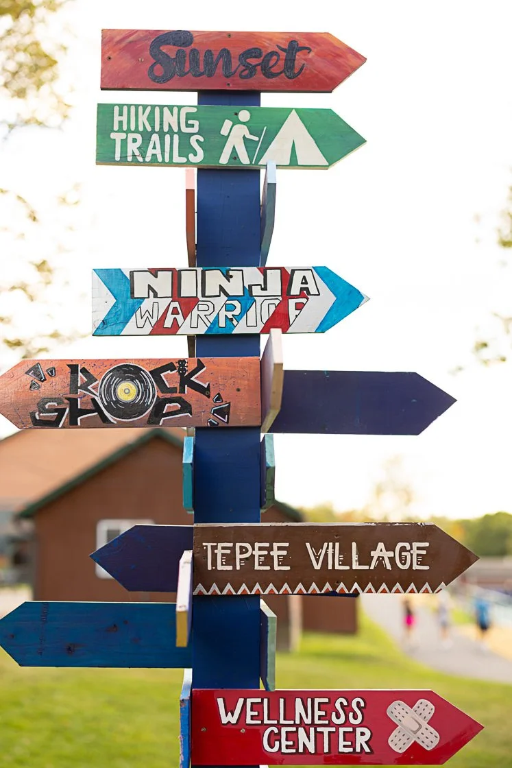 Colorful directional signs pointing to various activities and places, including Sunset, Hiking Trails, Ninja Warrior, Rock Shop, Tepee Village, and Wellness Center.