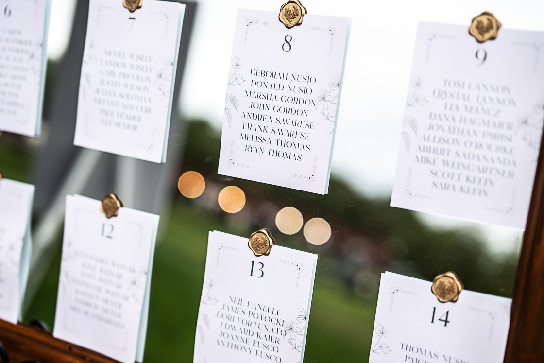 Wedding seating chart with white cards for table numbers and guest names, pinned on a glass surface with gold clips, blurred background with warm lights.
