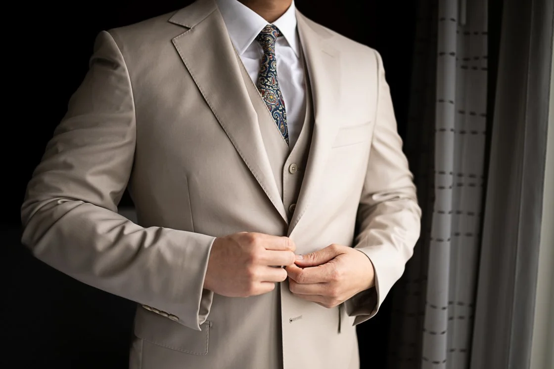 A man adjusting his beige suit jacket, wearing a white dress shirt and a patterned tie, standing by a window with curtains.