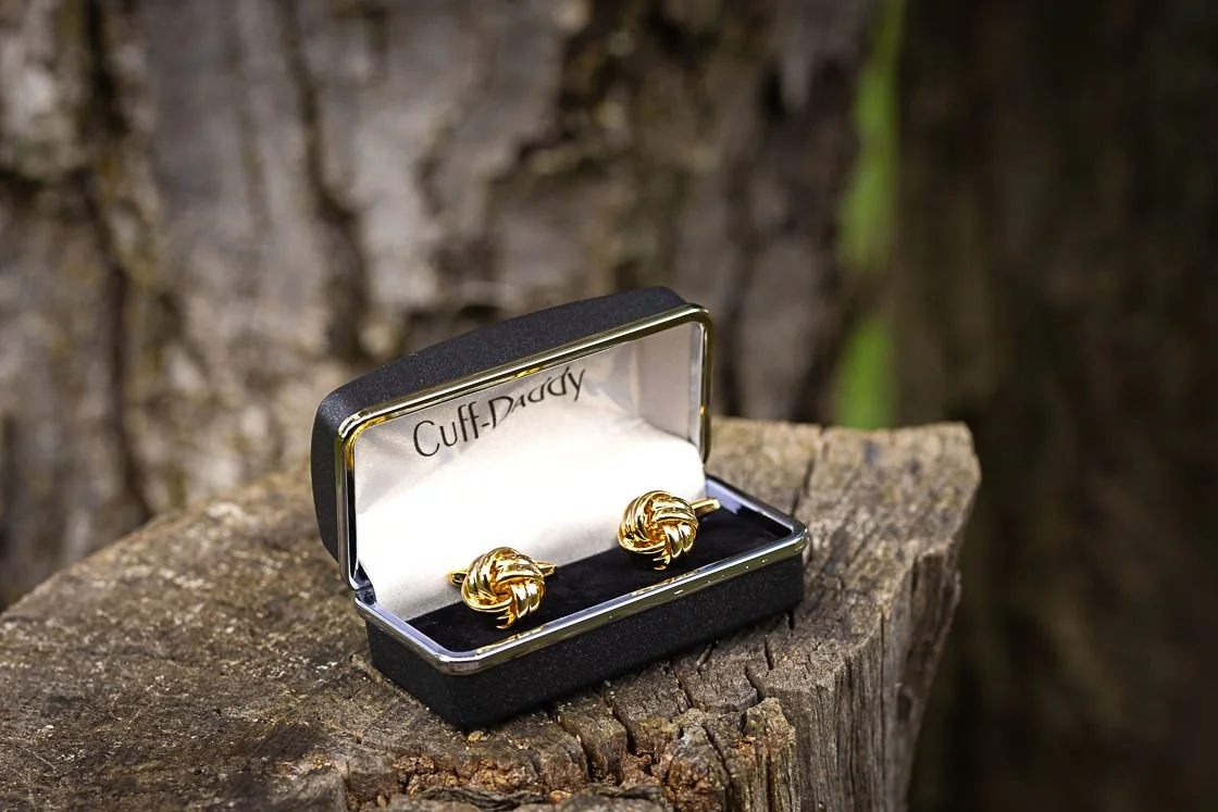 Gold knot cufflinks in a black jewelry box on a piece of wood with a tree bark background.