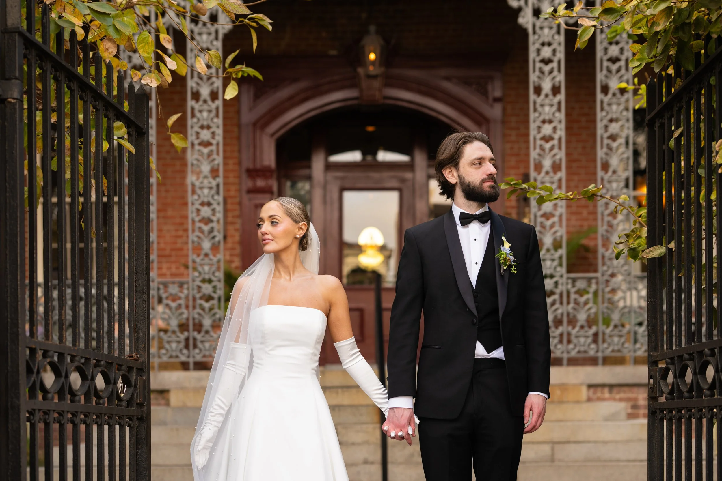 A bride and groom holding hands standing outdoors in front of a brick building with white decorative trim, with black iron gates framing the scene.