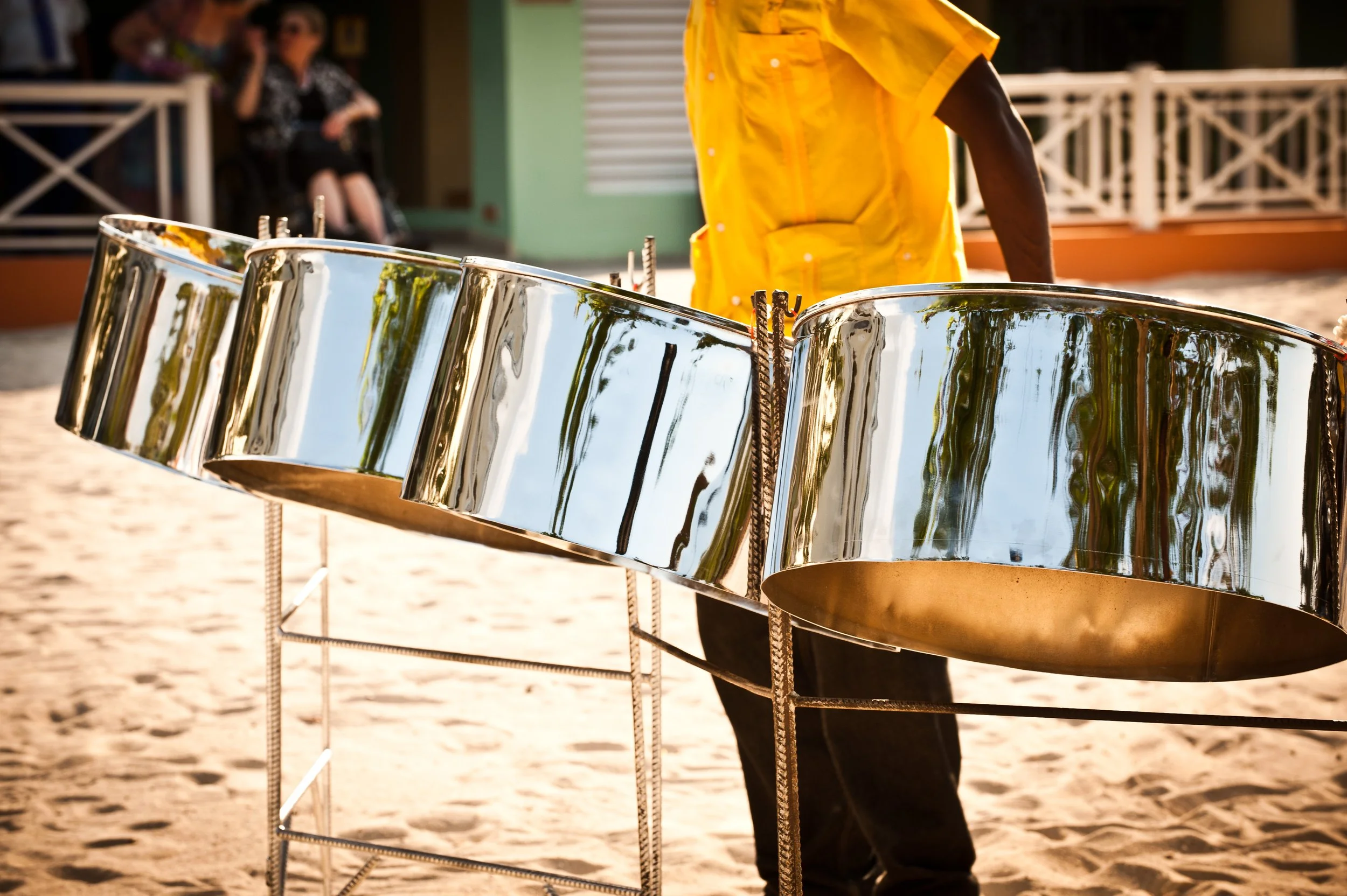 Person wearing a yellow shirt playing steel drums on a sandy beach while others watch nearby.