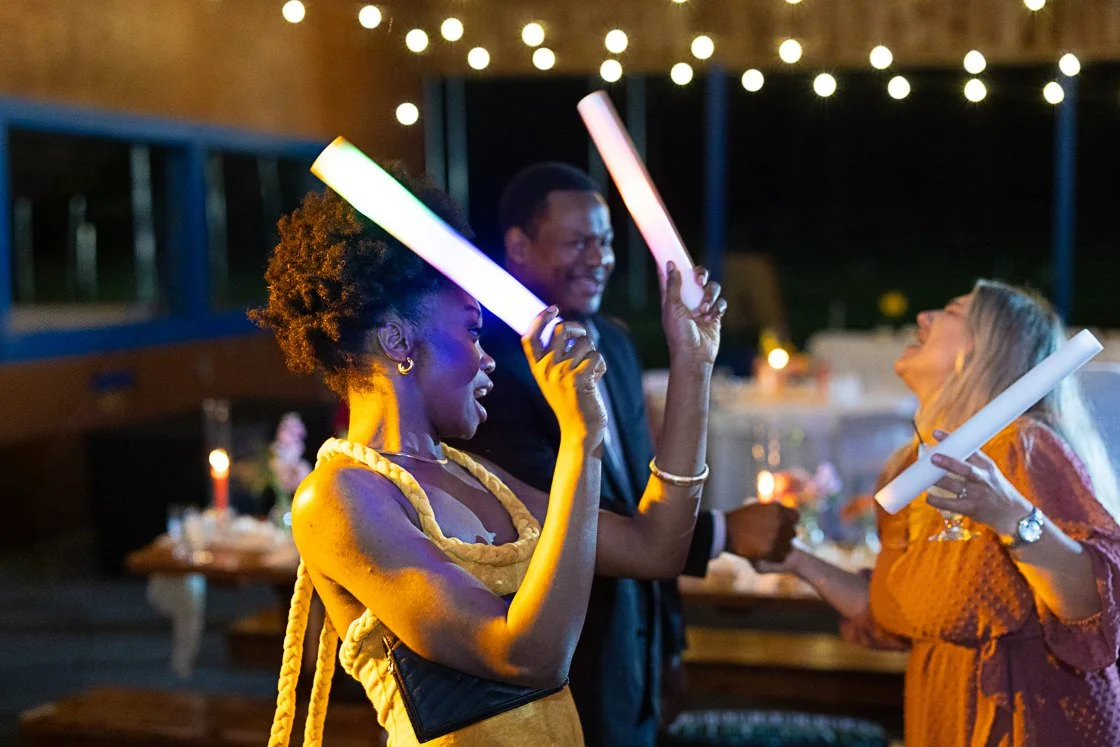 People celebrating at a party, holding glowing light sticks, with string lights overhead and a decorated table in the background.