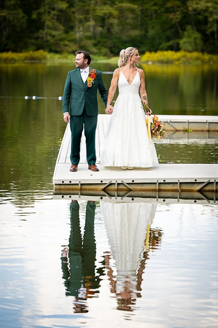 A bride and groom holding hands on a dock by a lake with forest background, reflected in the water.