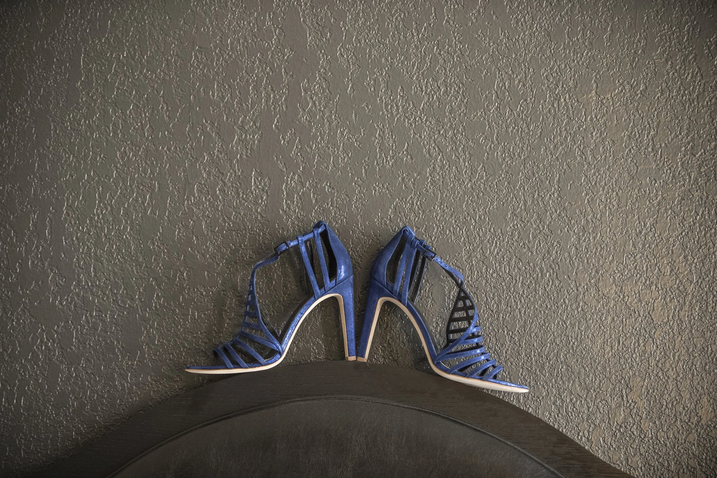 Pair of blue strappy high-heeled shoes on a textured gray floor, with part of a dark object and a person’s black clothing at the bottom edge of the image.