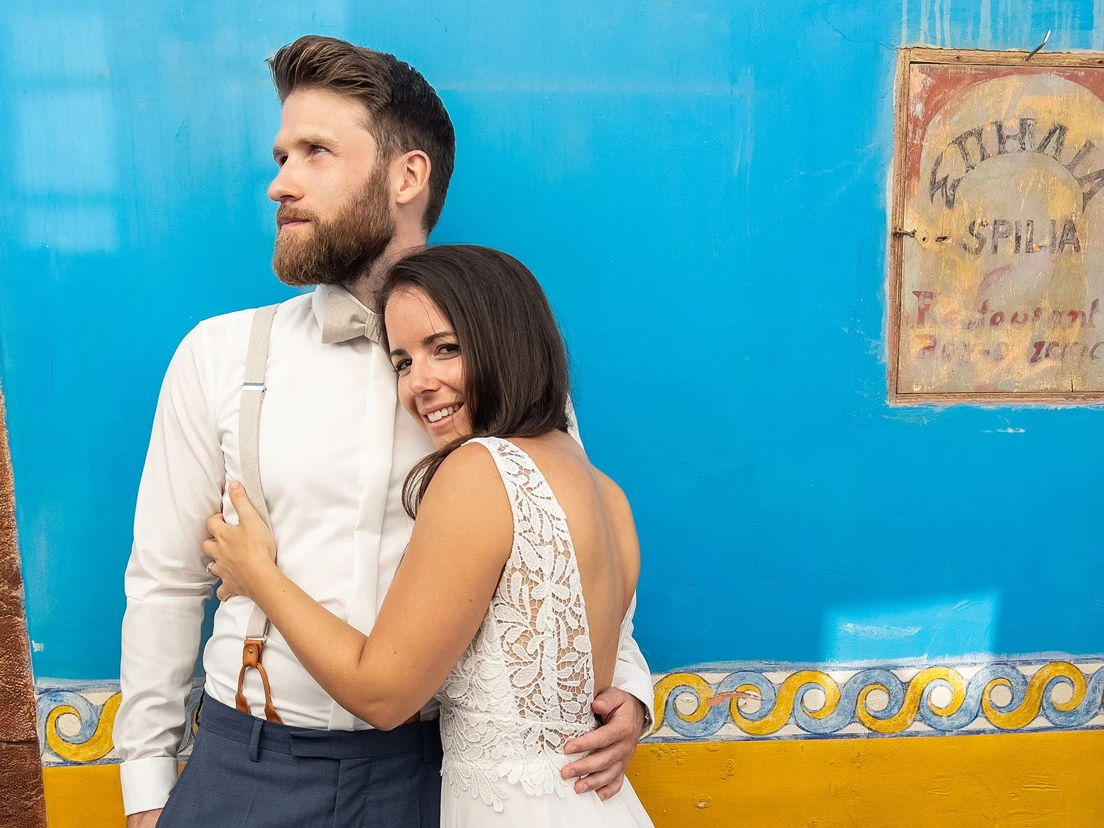 A happy couple standing in front of a bright blue wall with a decorative yellow and blue border, the woman smiling and hugging the man, both dressed in stylish clothing.
