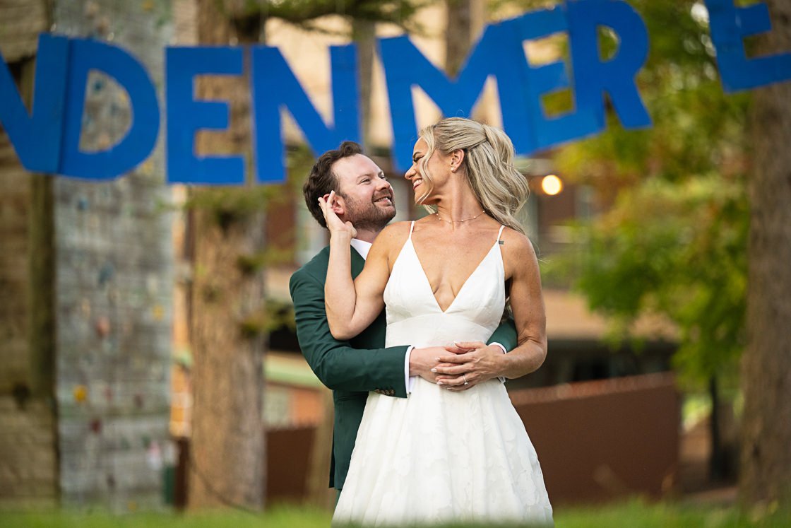 A couple in wedding attire celebrating outdoors, with a large blue 'DENGER' sign in the background, surrounded by trees and greenery.