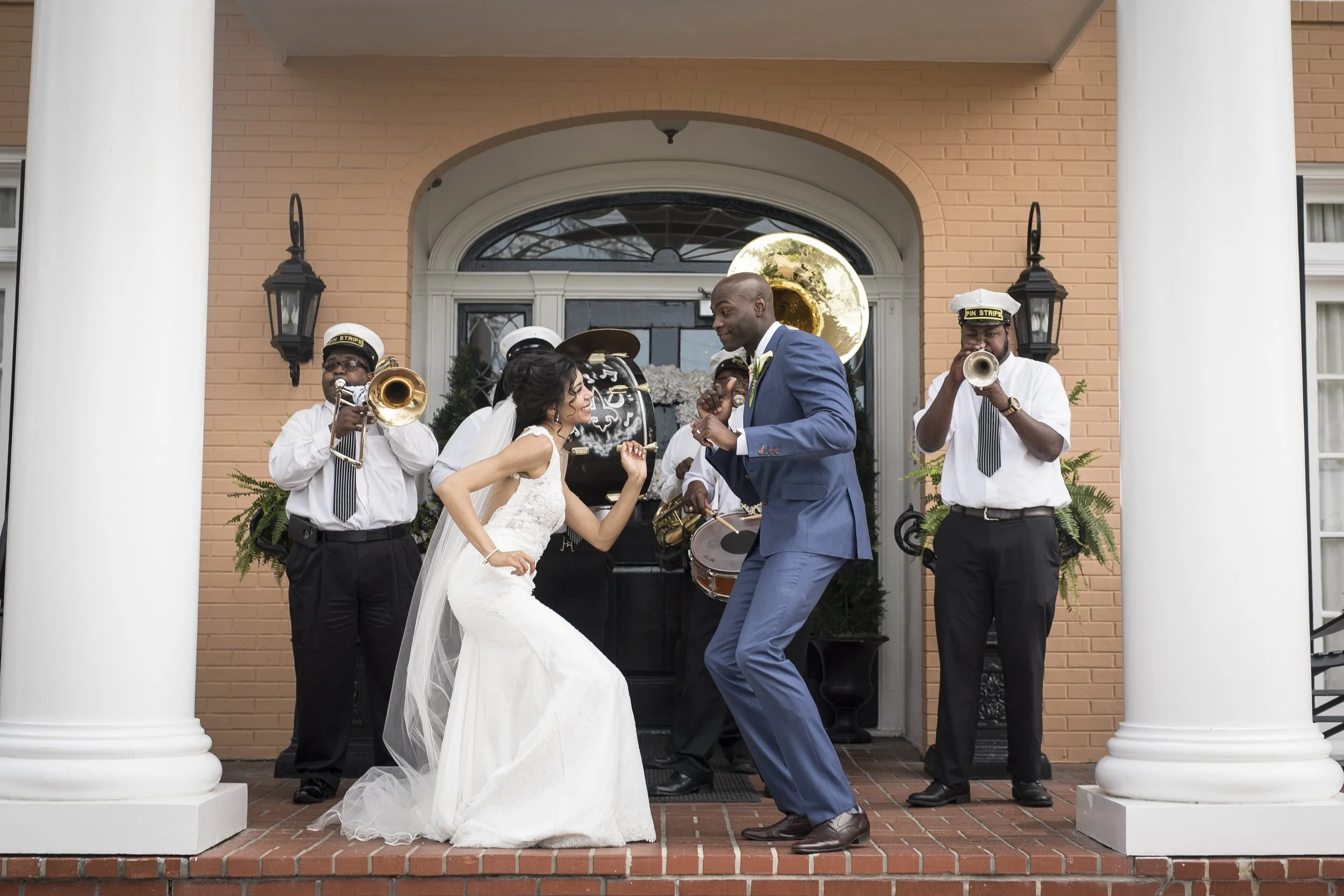 A bride and groom dancing on the steps of a building with a brass band playing behind them. The building has orange brick walls, white columns, and is decorated for a wedding.