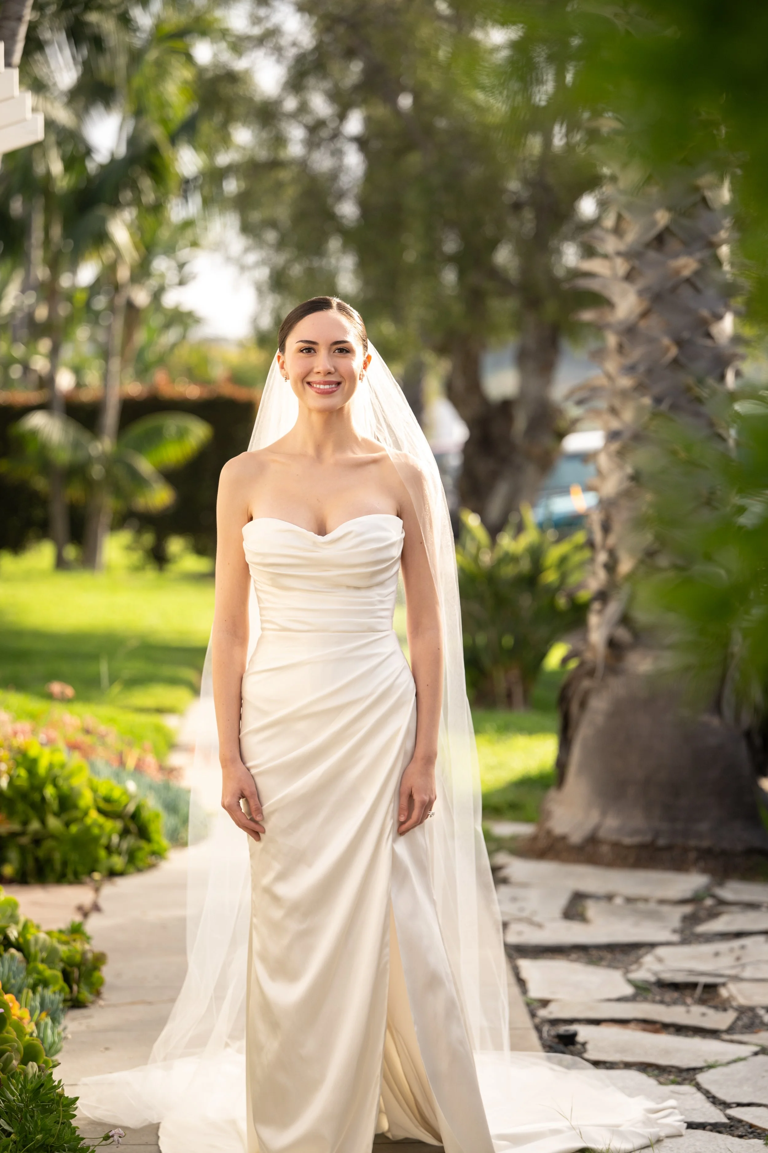A smiling bride in a strapless white wedding gown and veil standing on a garden pathway surrounded by greenery and palm trees.