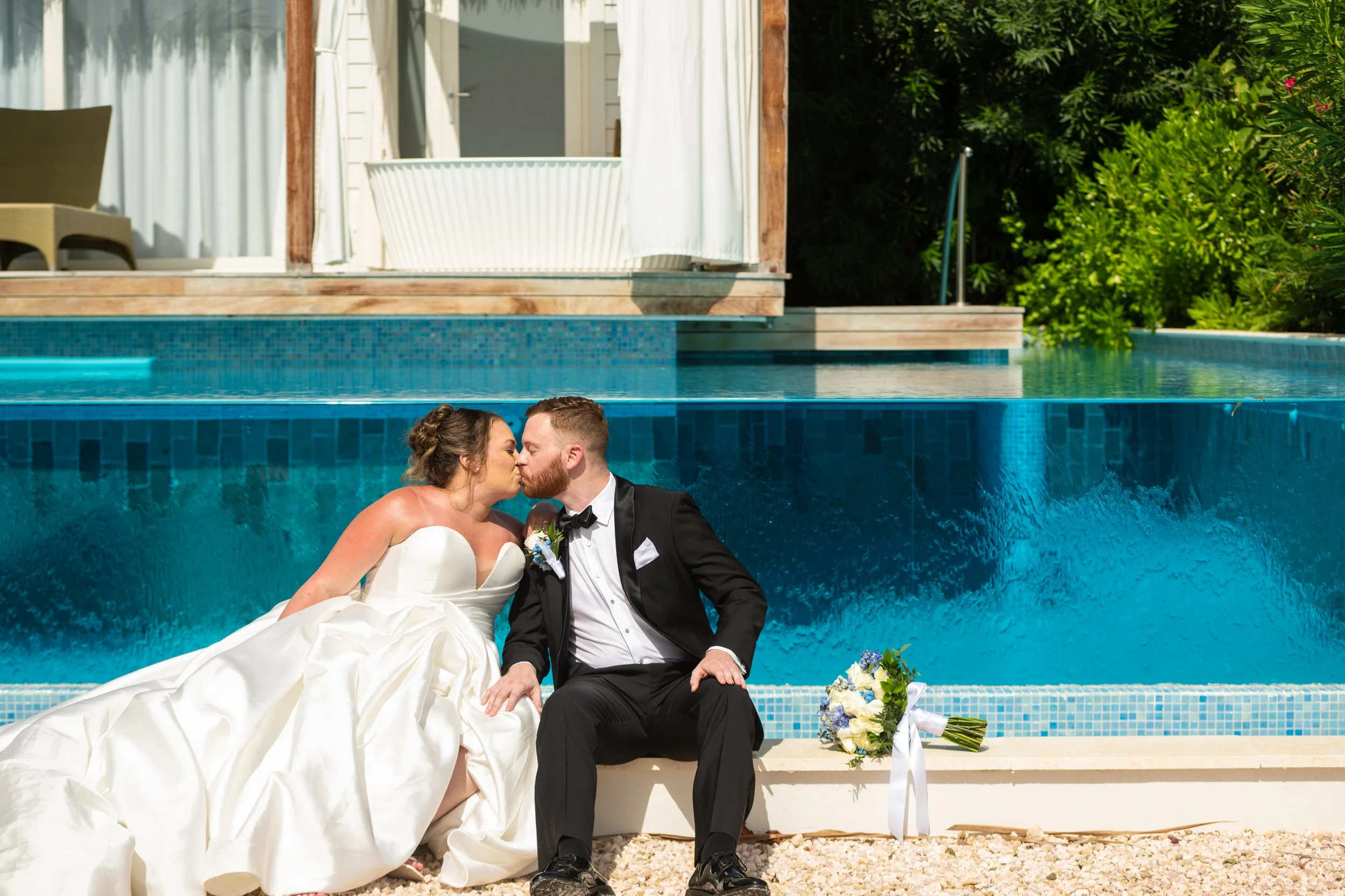 A bride and groom sit together by a pool, sharing a kiss after their wedding, with a bouquet of flowers nearby and greenery in the background.