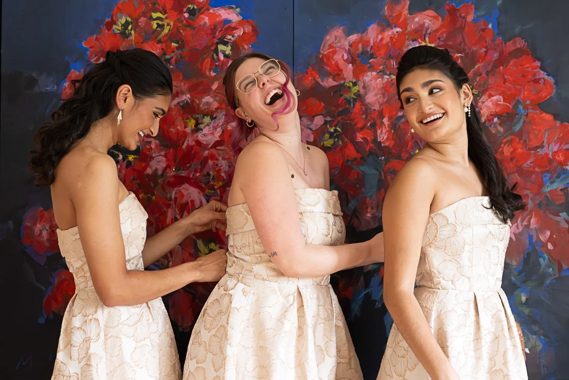 Three women in matching cream-colored strapless dresses laughing and holding hands in front of a vibrant red floral background.
