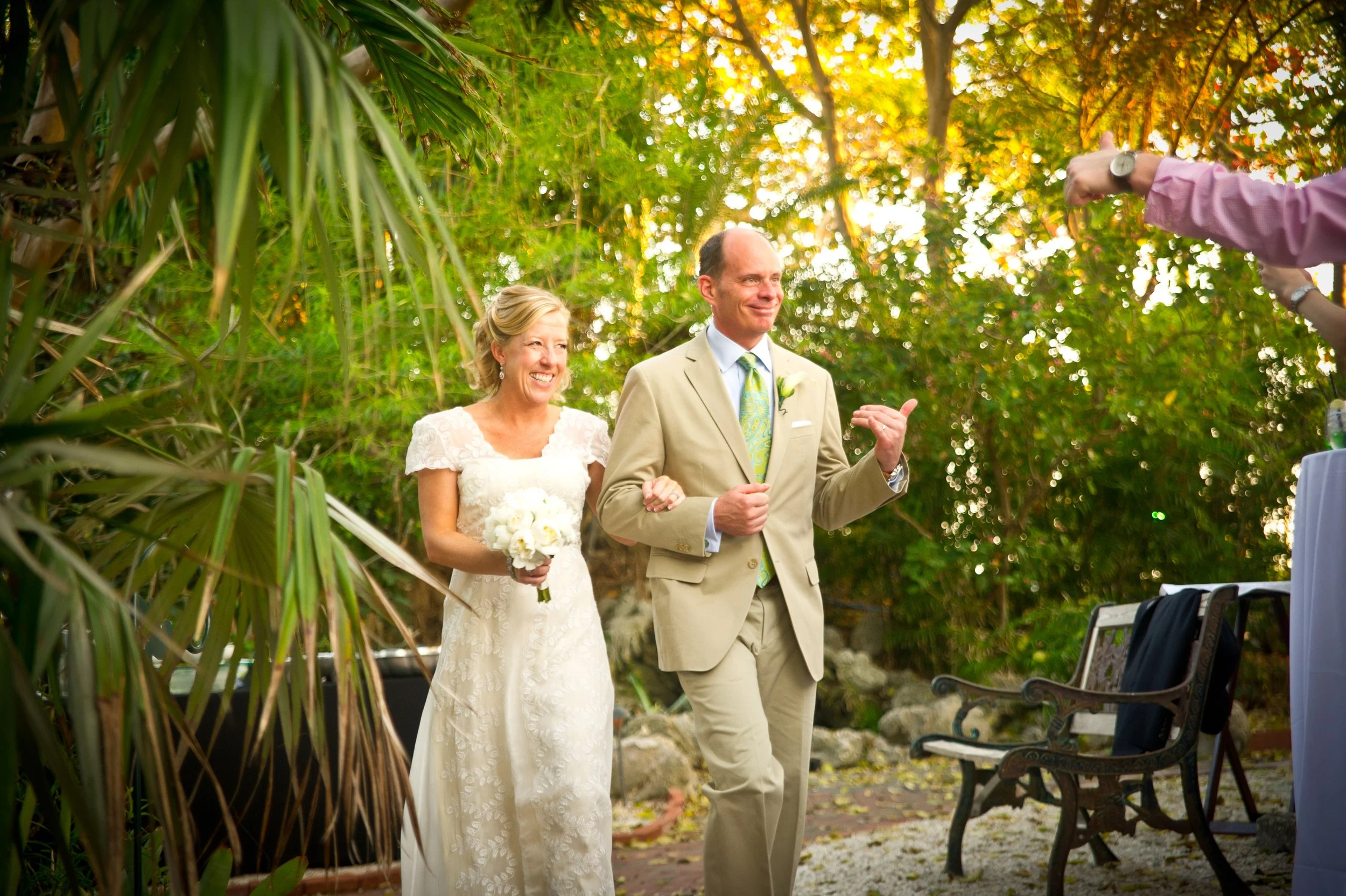 A bride in a white lace wedding dress holding a bouquet of white flowers and a groom in a beige suit walking outdoors during a wedding ceremony. They are smiling, and the groom gestures with a thumb up. The setting features lush greenery and trees wi
