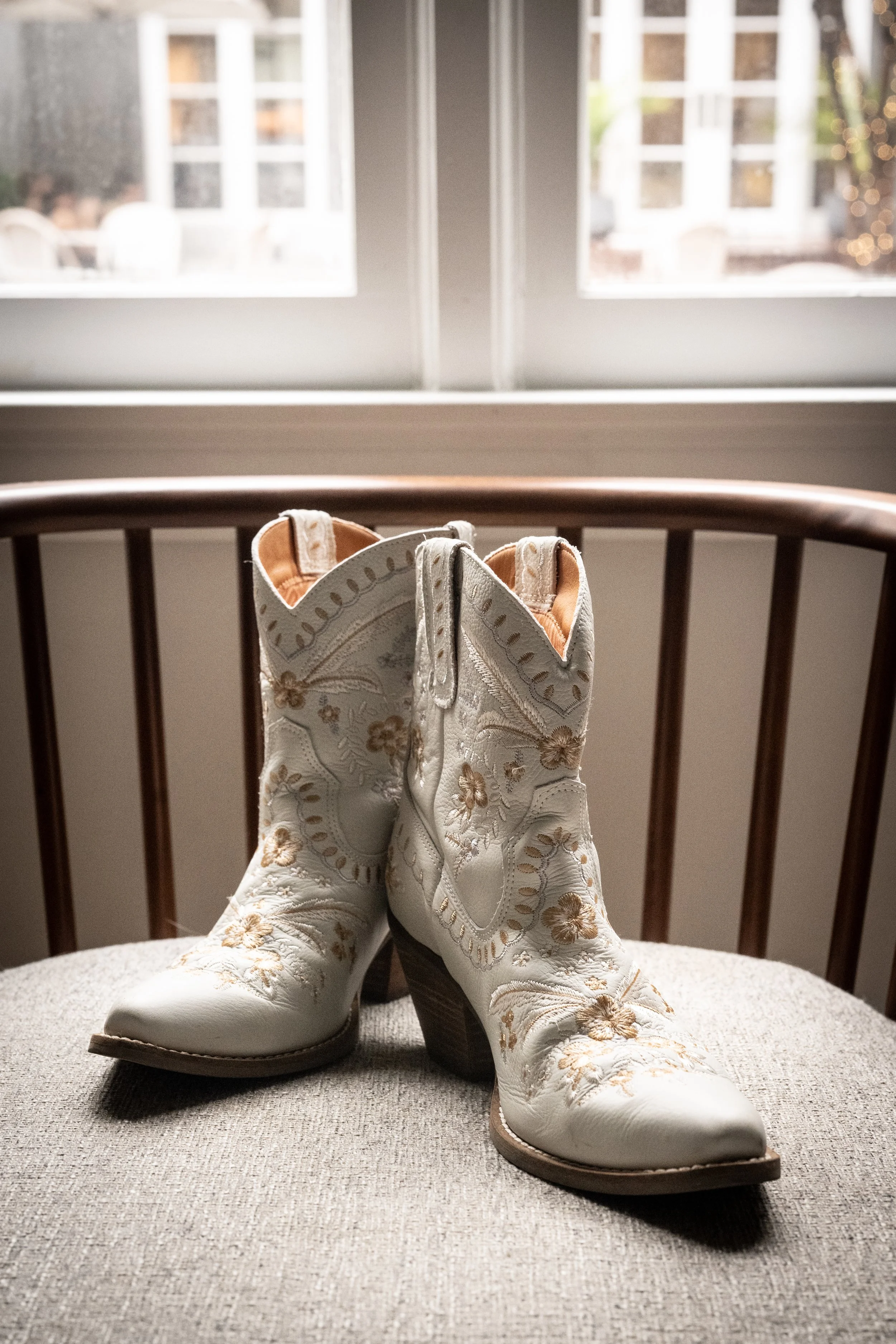 Pair of white cowboy boots with floral embroidery, resting on a cushioned surface in front of a wooden chair and a window with a garden view.