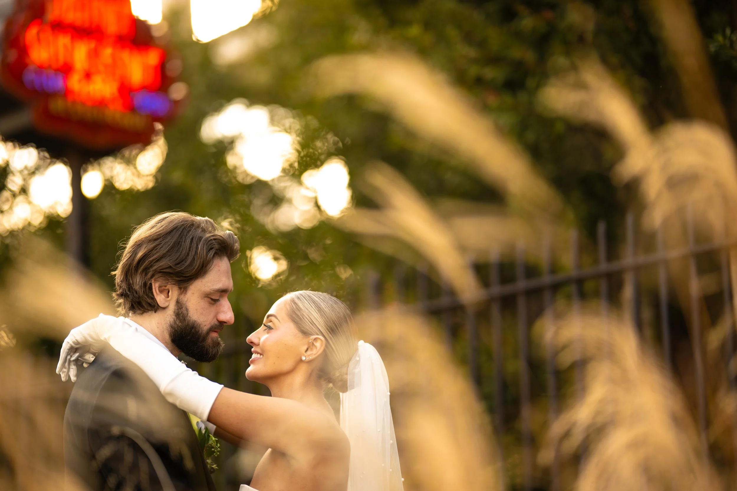 A close-up of a bride and groom facing each other and smiling, with the groom having dark hair and a beard, the bride with blonde hair and earrings, on a sunny outdoor setting with blurred trees and a fence in the background.