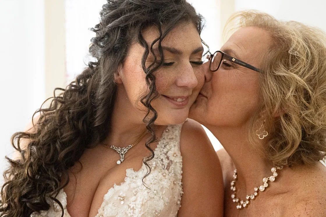 A woman in a wedding dress receiving a kiss on the cheek from another woman wearing glasses and jewelry.