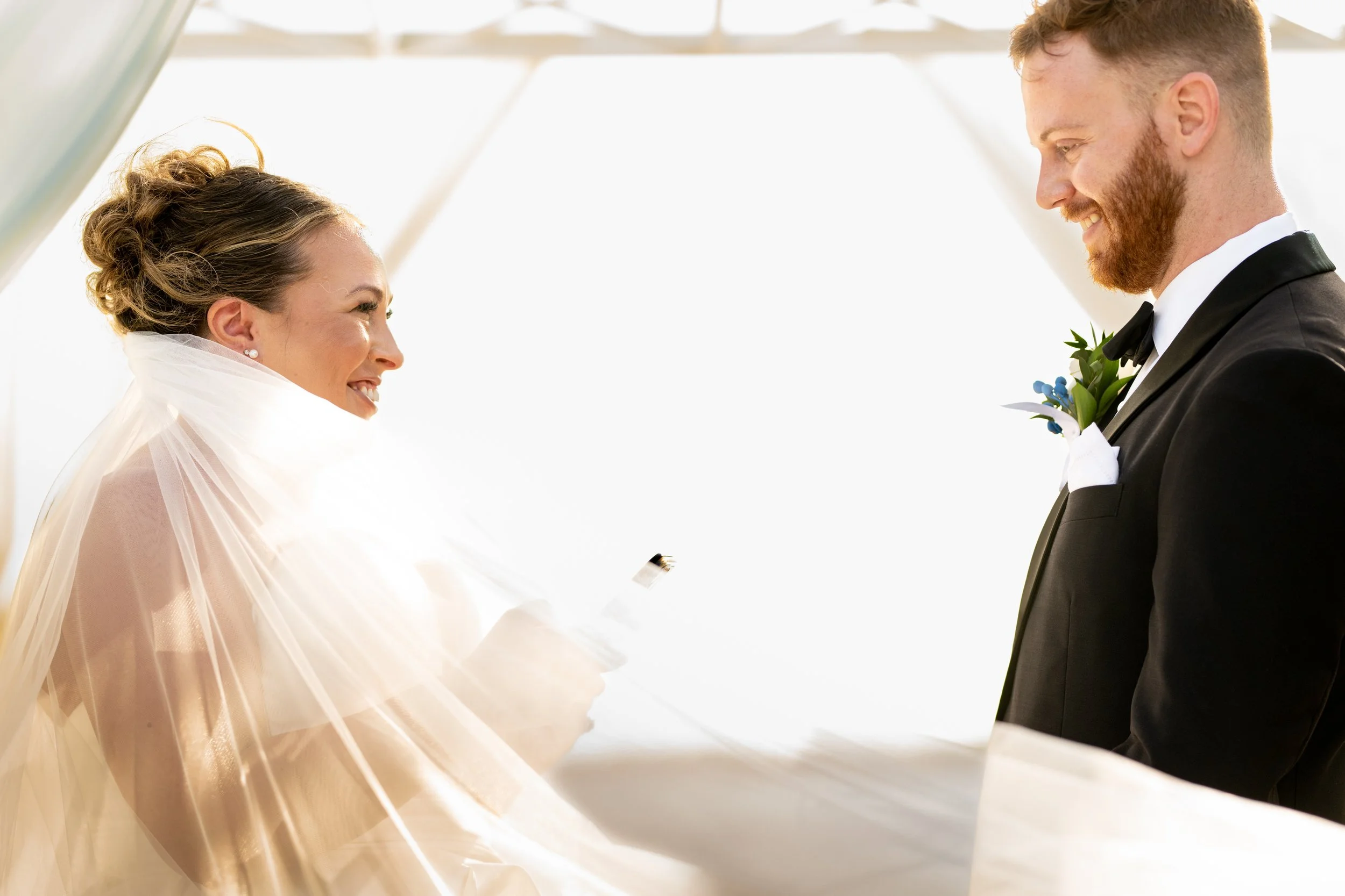 A bride and groom smiling at each other during a wedding ceremony, with the bride holding a ring box.