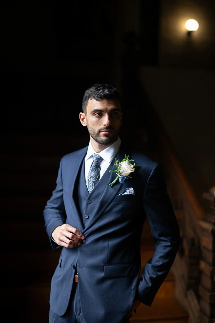 A man in a navy suit with a white shirt and patterned tie, wearing a boutonnière, standing indoors with a dim background and soft lighting.