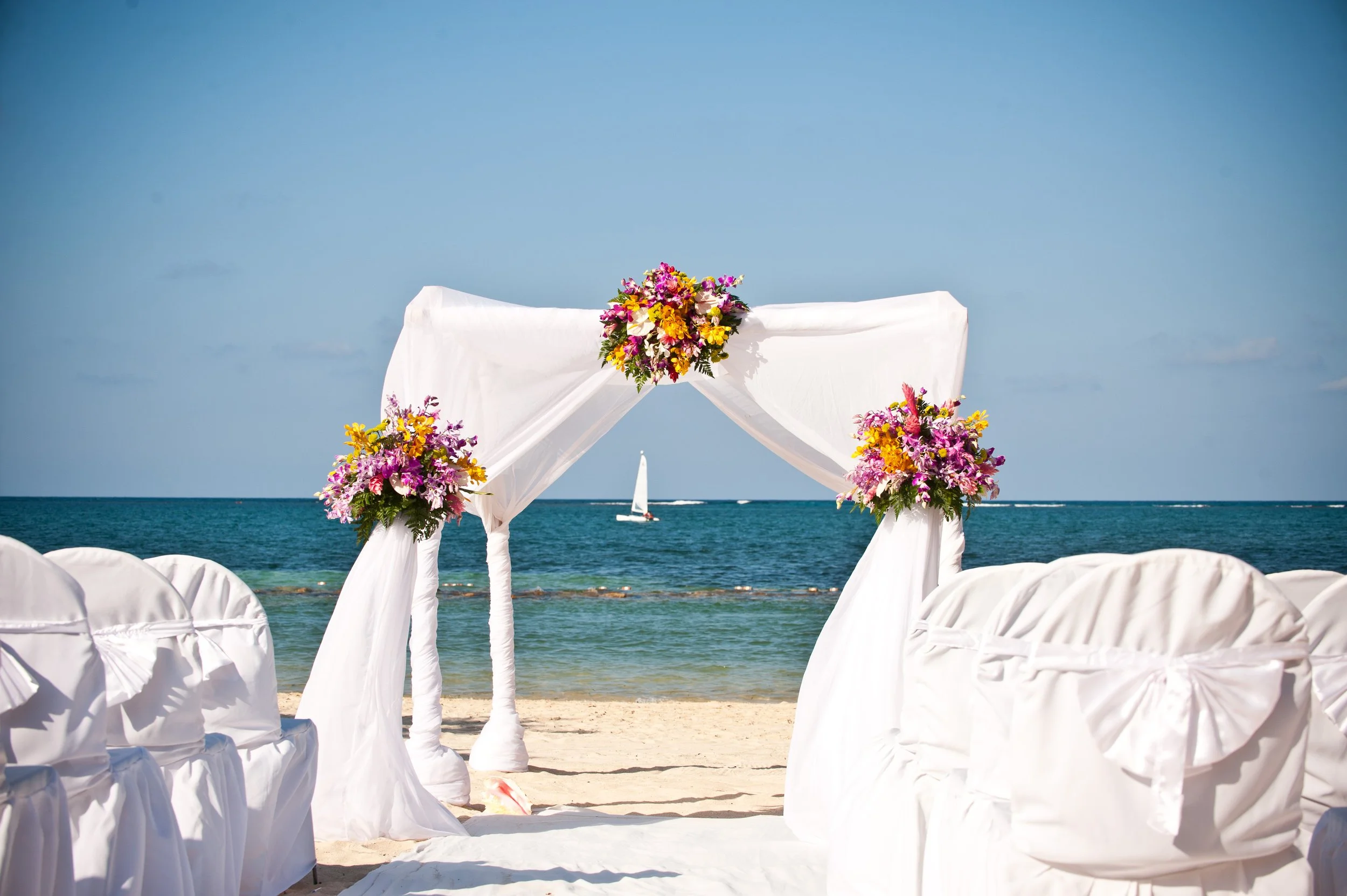A wedding arch with white fabric and colorful flowers set up on a beach, with chairs draped in white fabric facing the arch, and boats sailing on the ocean in the background.