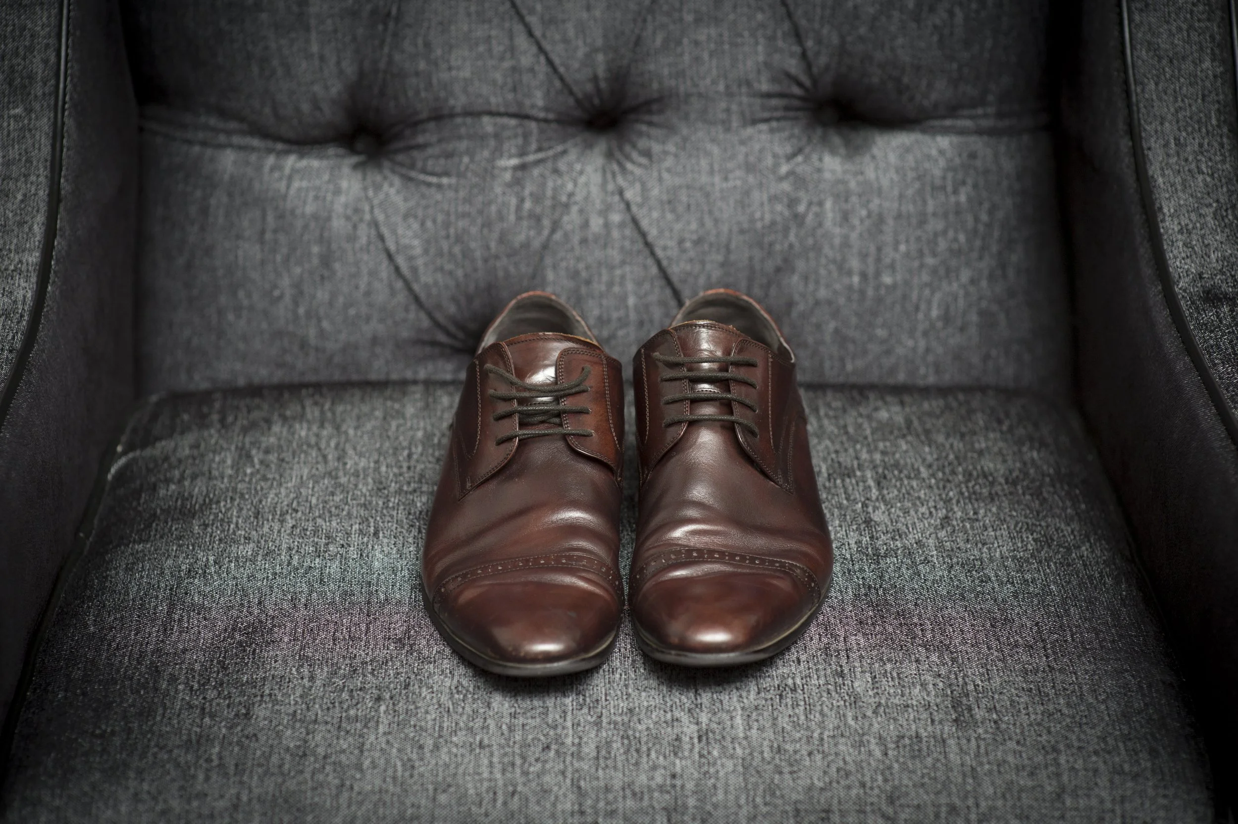 A pair of brown leather dress shoes on a gray upholstered armchair.