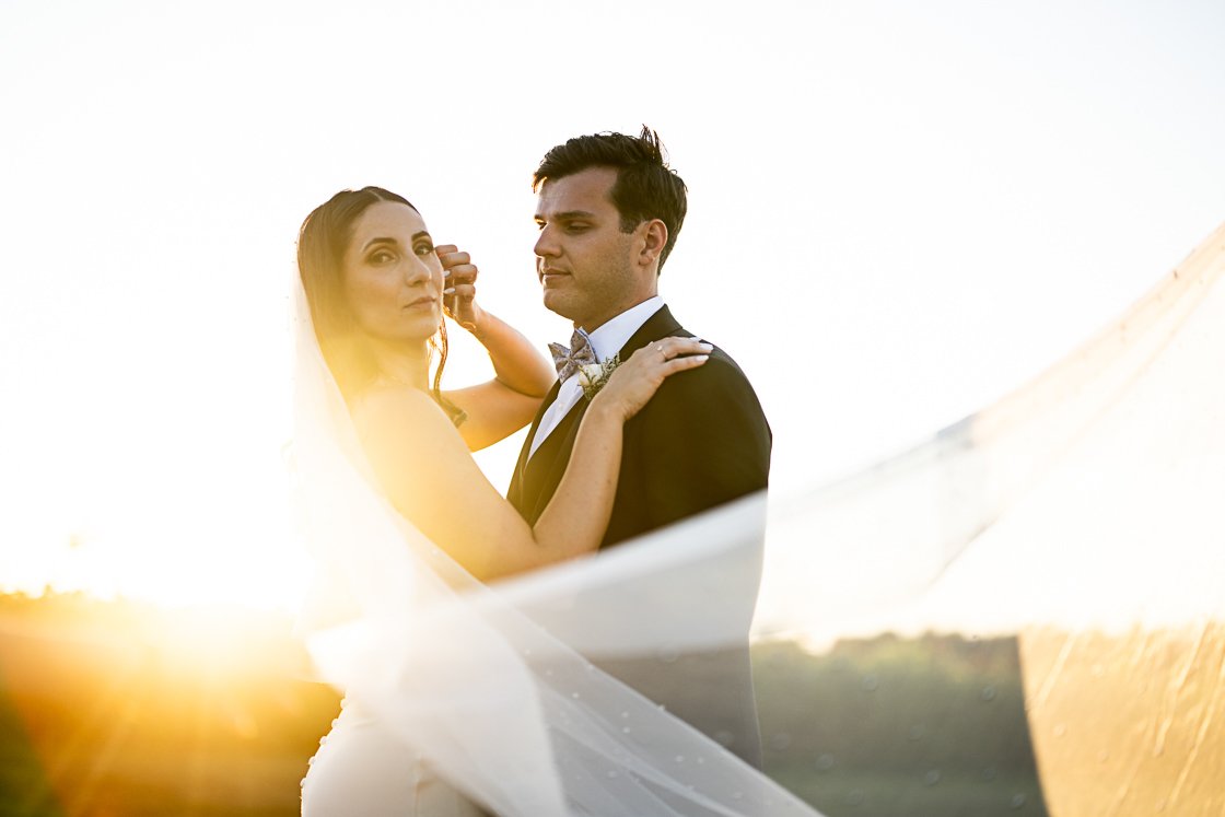 A bride and groom in wedding attire, embracing outdoors at sunset, with the bride's veil flowing in the breeze.