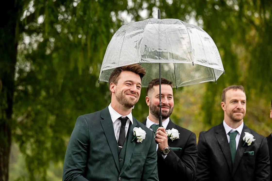 Three men dressed in suits, one holding a transparent umbrella over two of them during a rainy outdoor event
