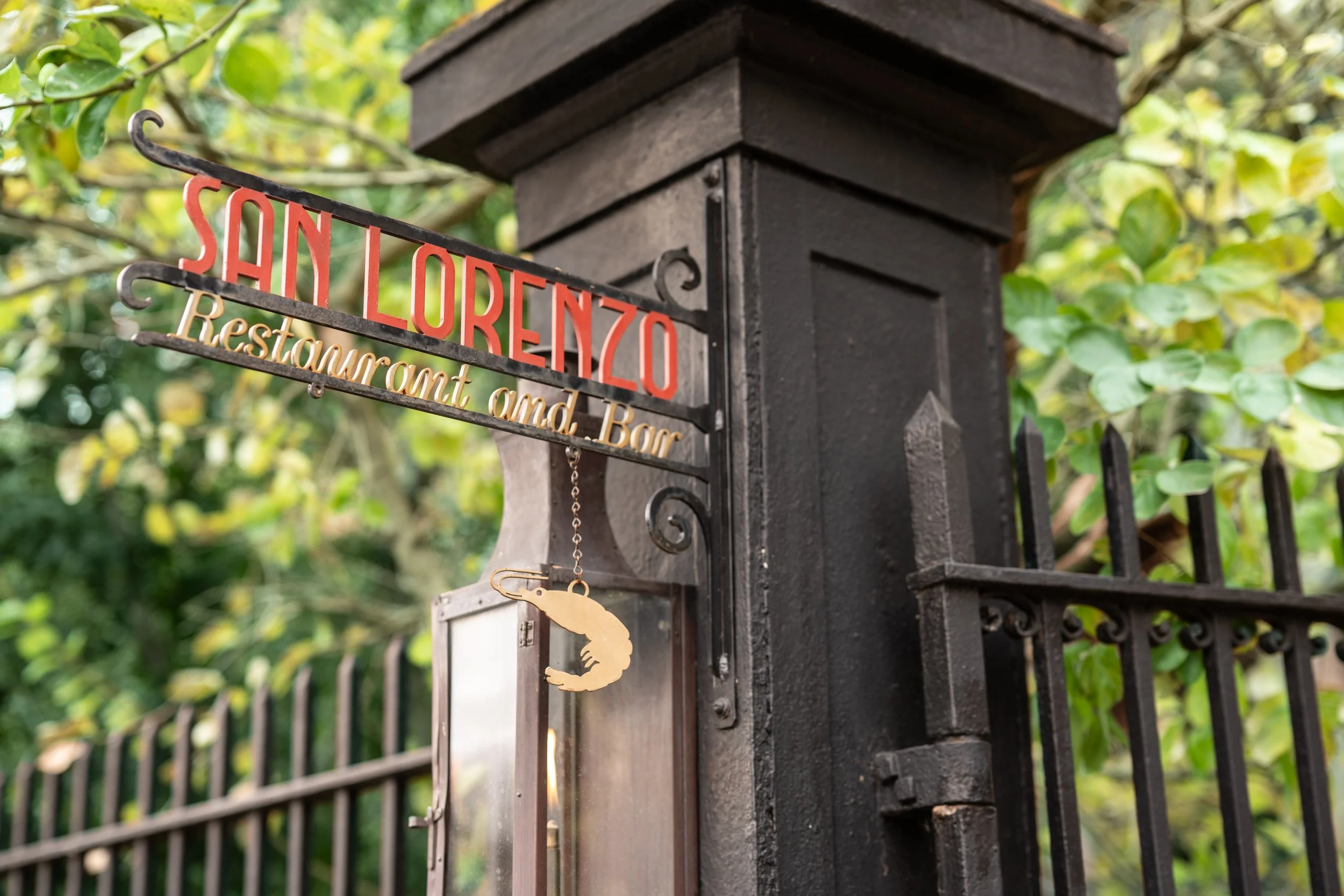 A black metal mailbox with a wooden post and a sign that reads 'SH LORENZO Restaurant and Bar' mounted on the post. Greenery in the background.