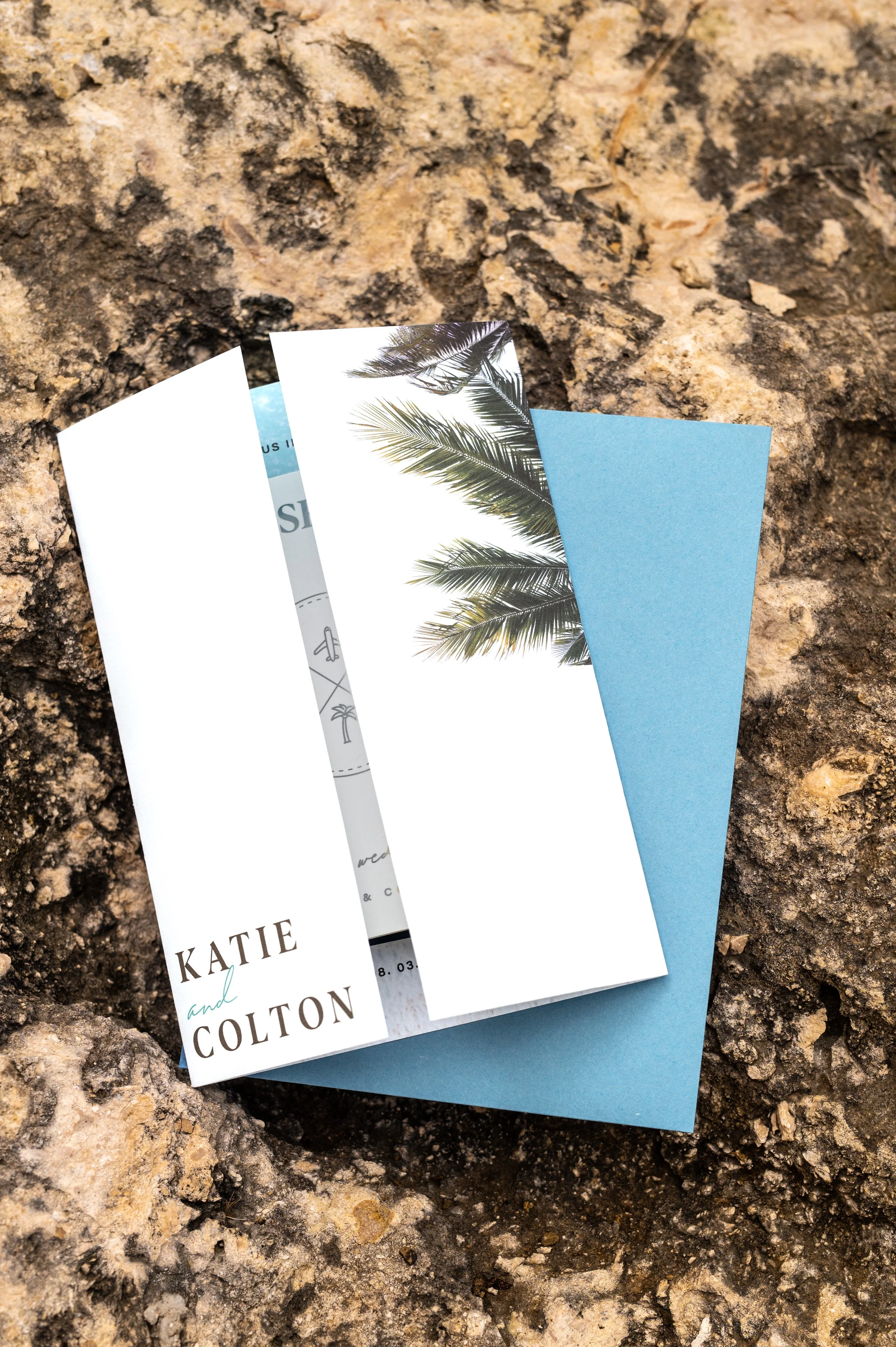 A wedding invitation with palm tree illustrations on a blue background, placed on a rough, rocky surface.