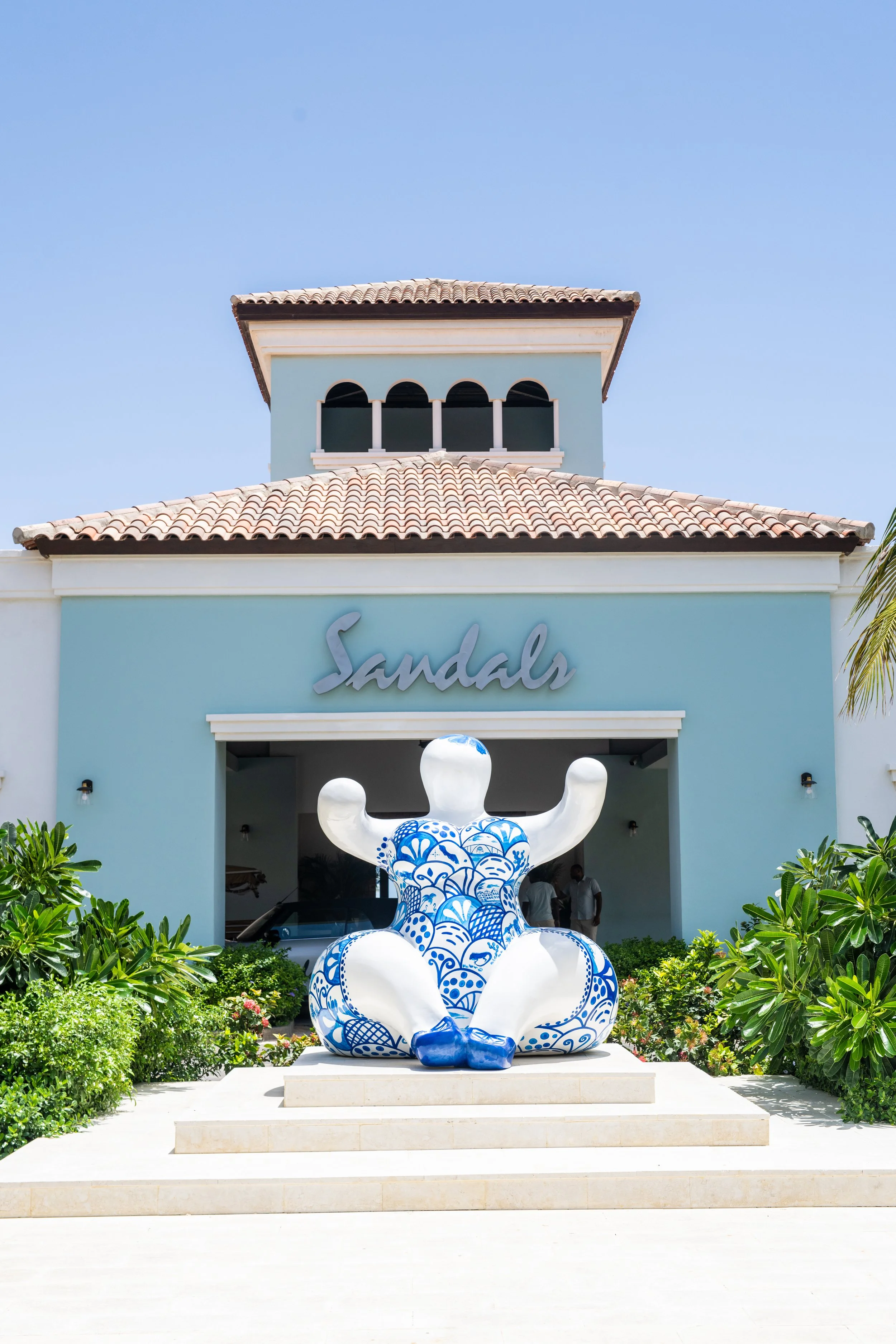 Exterior of Sandals resort with a large decorative blue and white lotus-shaped sculpture at the entrance, surrounded by green shrubs and plants, under a clear blue sky.