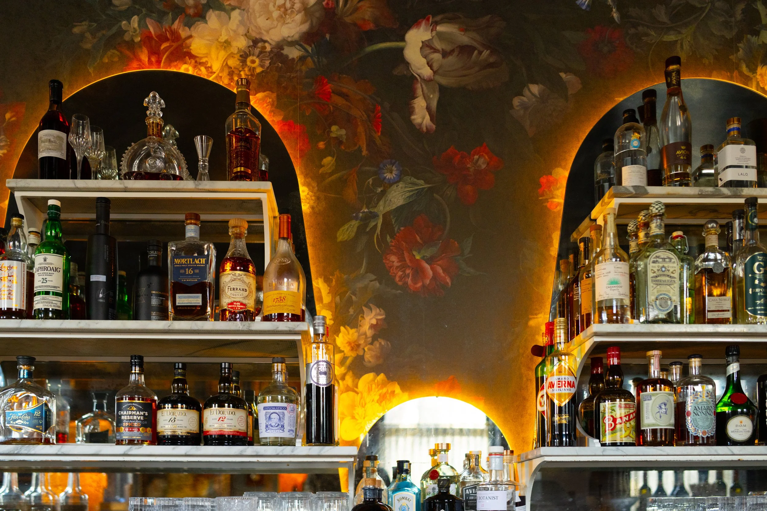 Bar shelves filled with various liquor bottles in front of a dark wall decorated with floral paintings and illuminated with warm lighting.