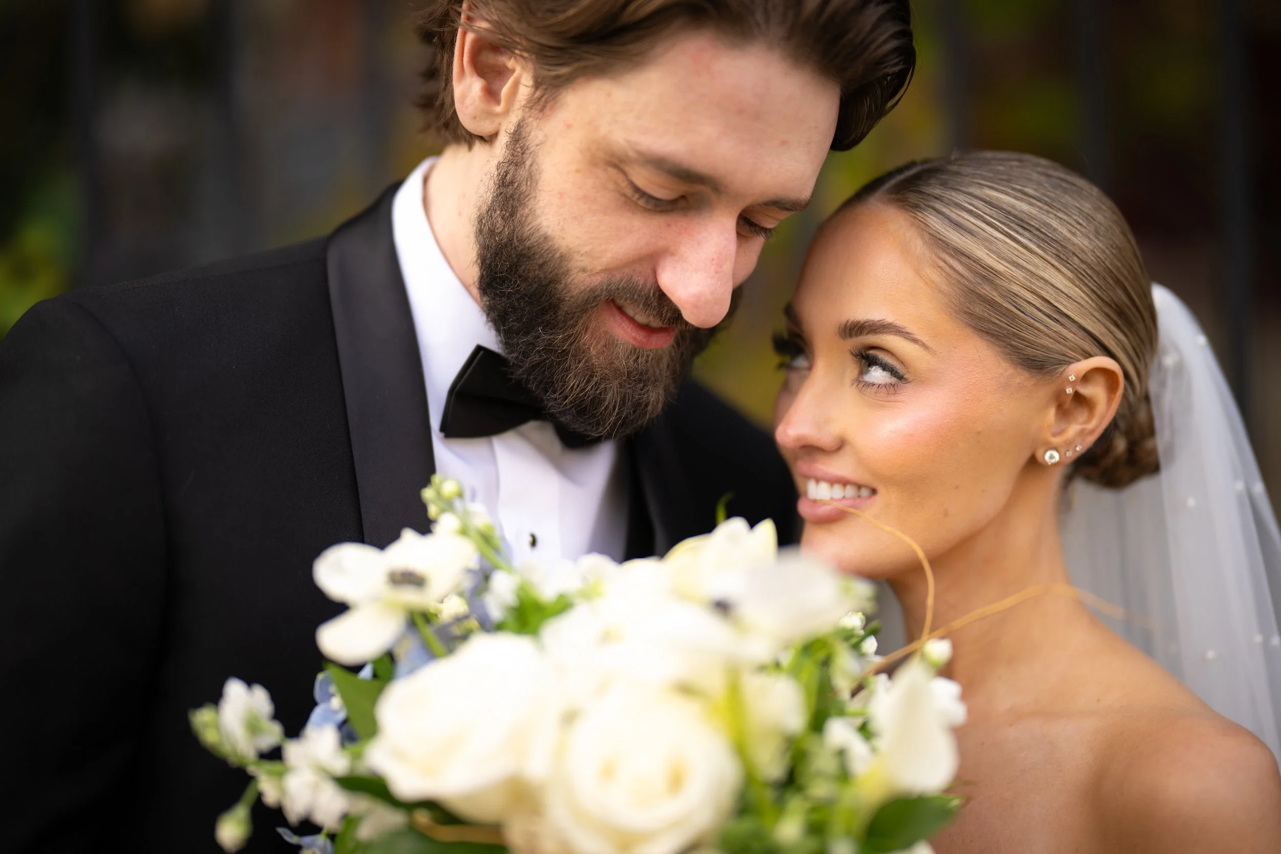 A close-up of a bride and groom smiling and leaning their foreheads together during a wedding, with the bride holding a bouquet of white flowers.