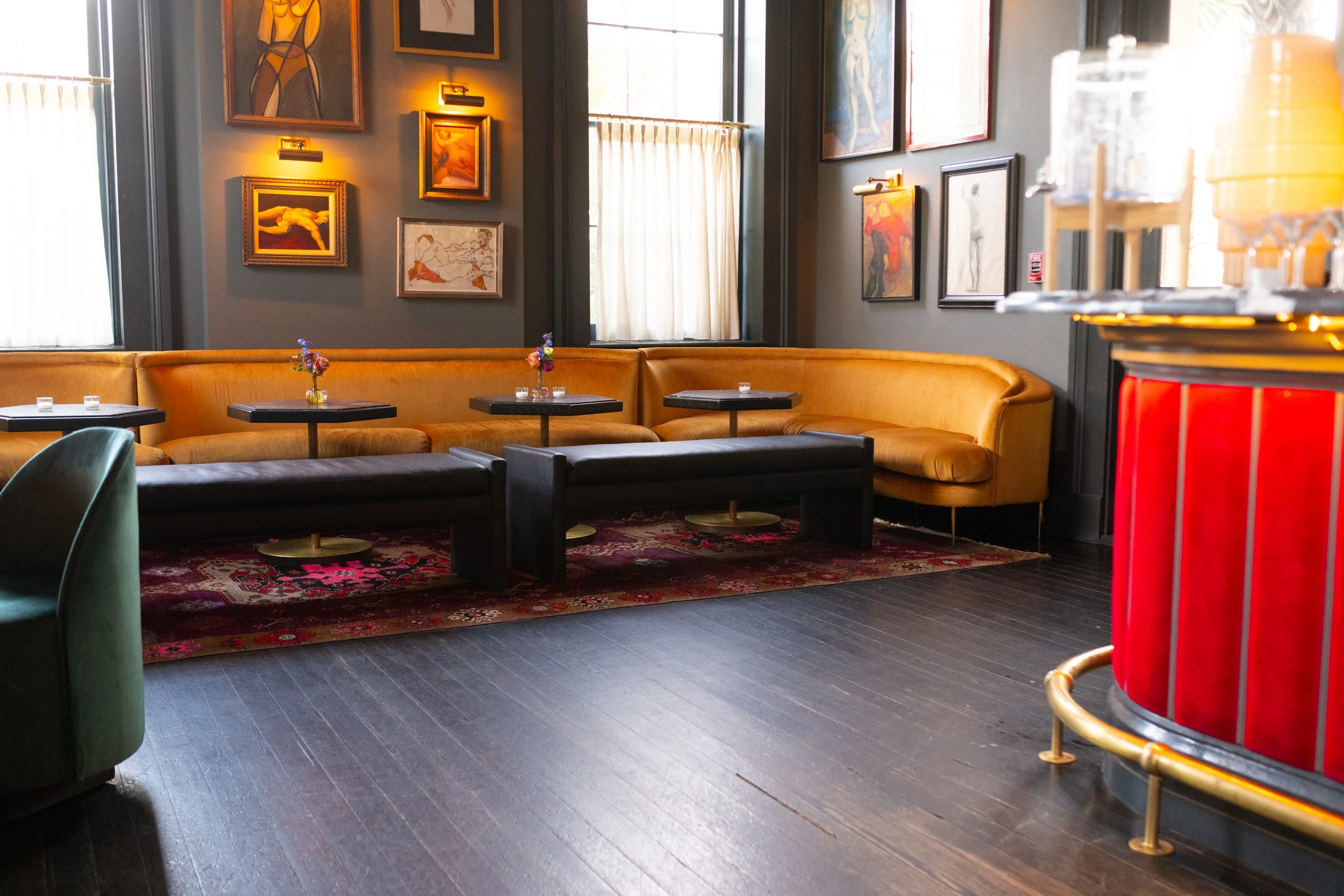 A cozy lounge area with yellowish-orange curved sofa, small black tables with vases of flowers, framed artwork on dark gray walls, window with sheer curtains, on a dark wooden floor with a patterned rug.