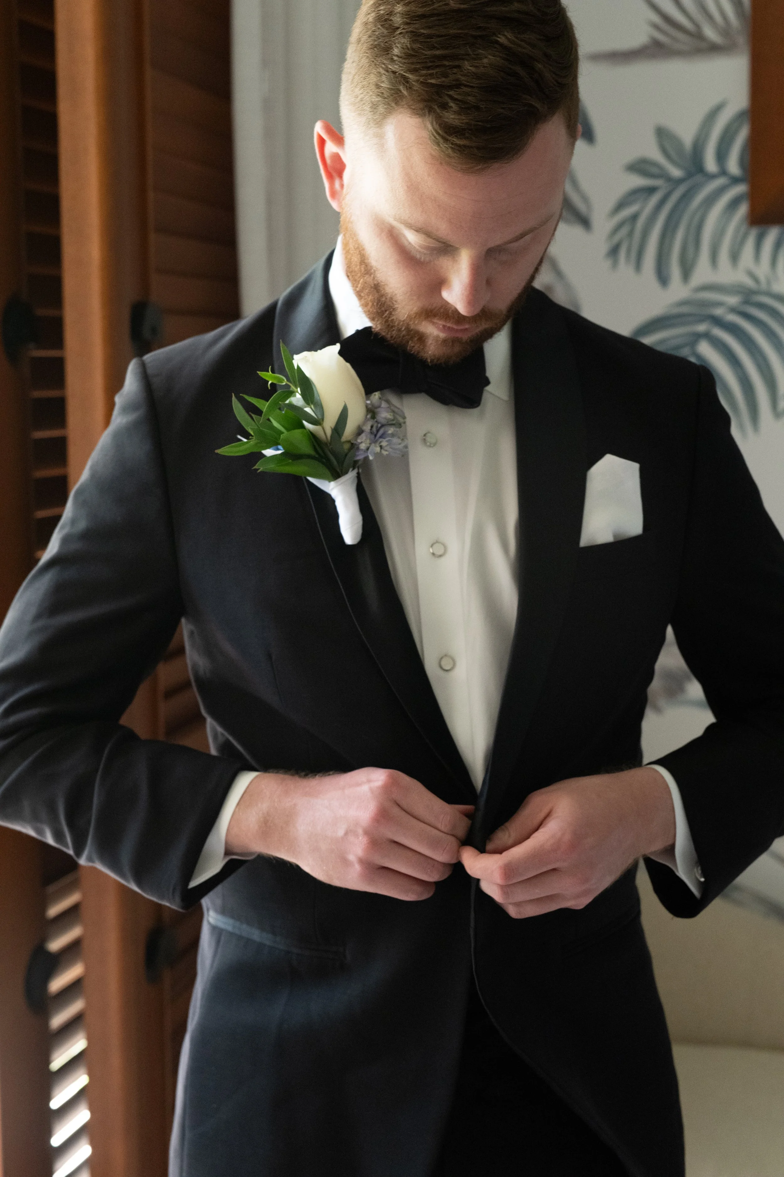 A man in a black tuxedo with a white shirt and black bow tie, adjusting his jacket. He has short, light brown hair and a beard. He is wearing a white boutonniere on his left lapel. The background features wooden furniture and a wall with a leaf patte