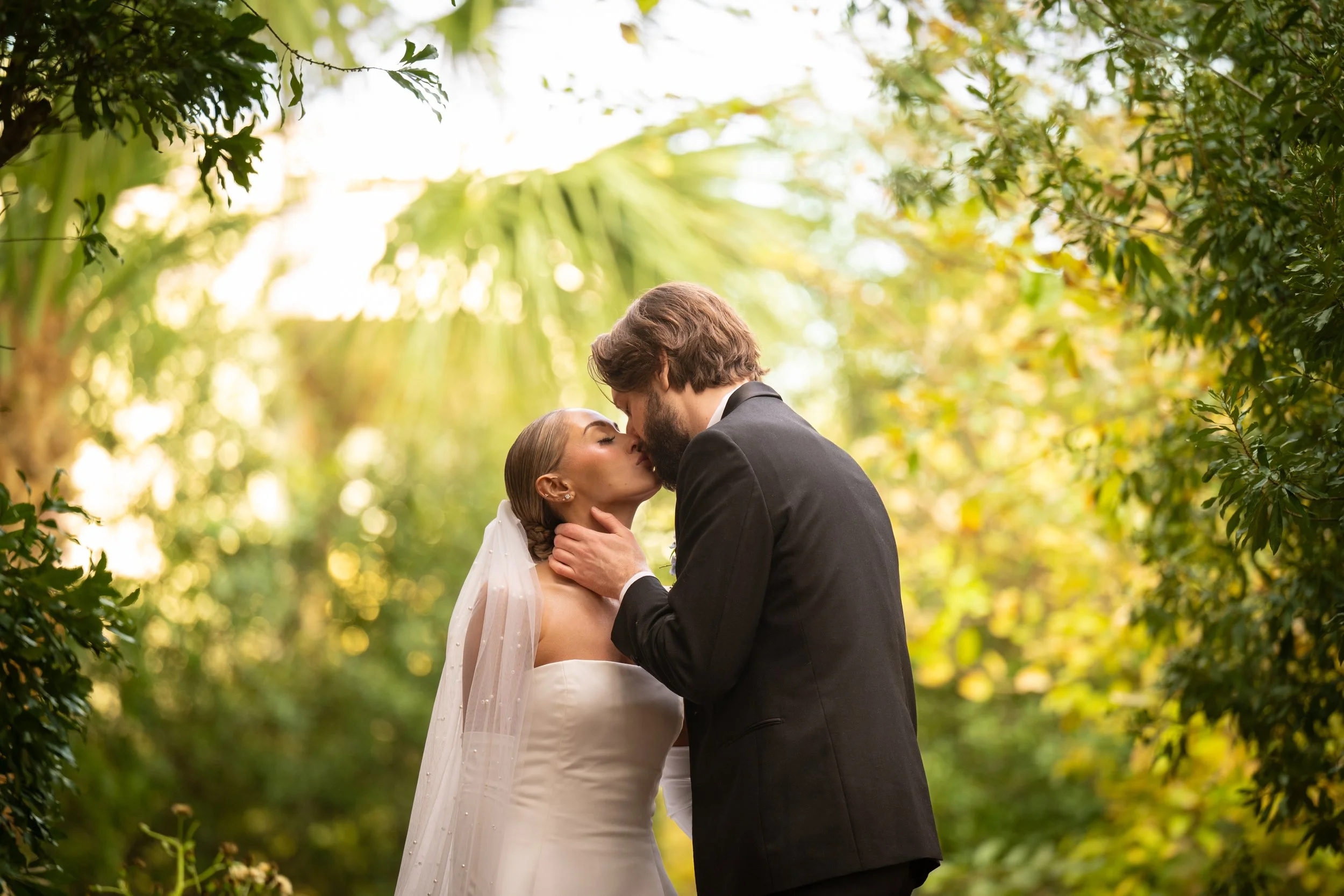 A bride and groom share a kiss outdoors in a lush green, sunlit setting during their wedding.