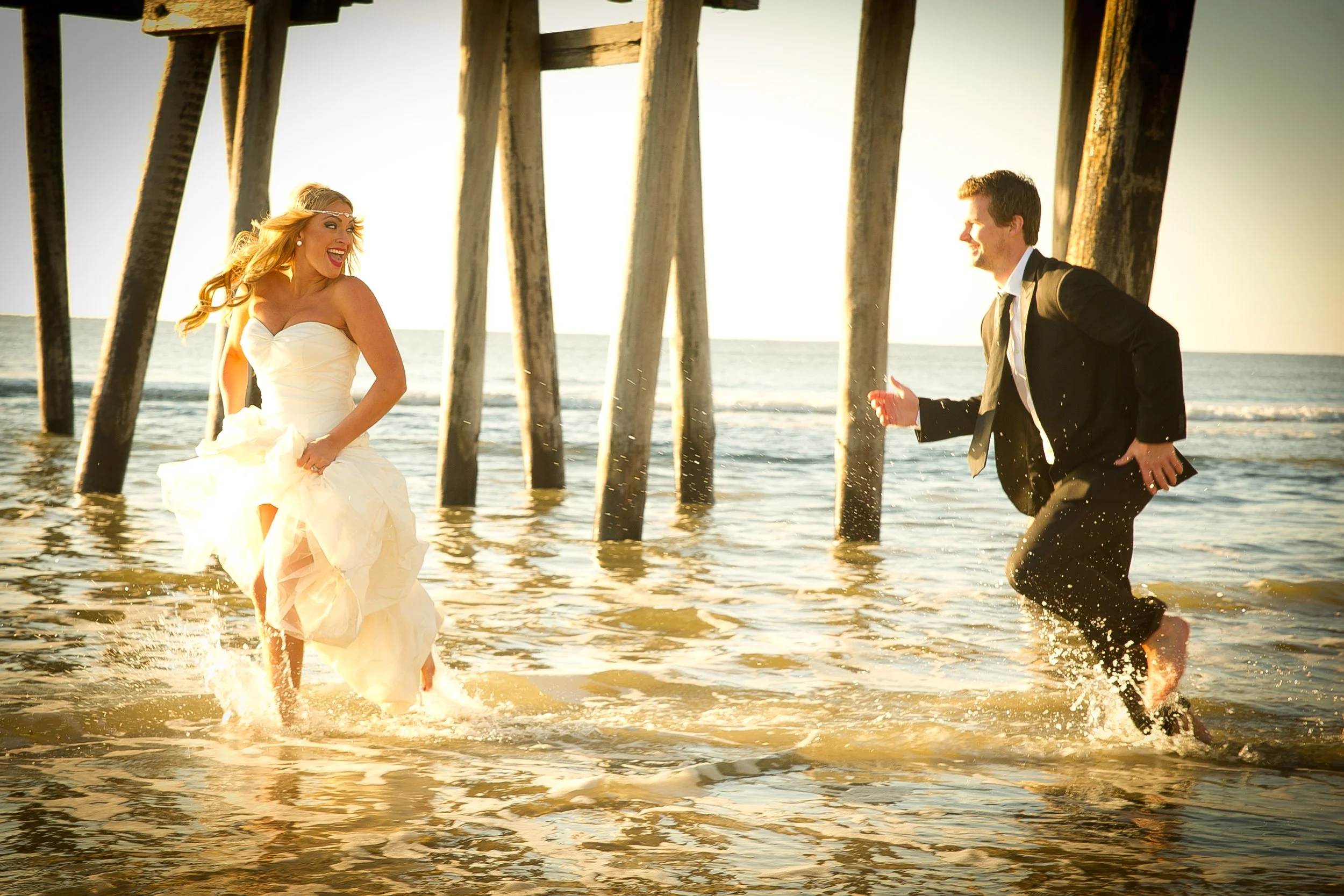 A bride and groom running in shallow water under a pier on the beach at sunset, smiling and enjoying their wedding.