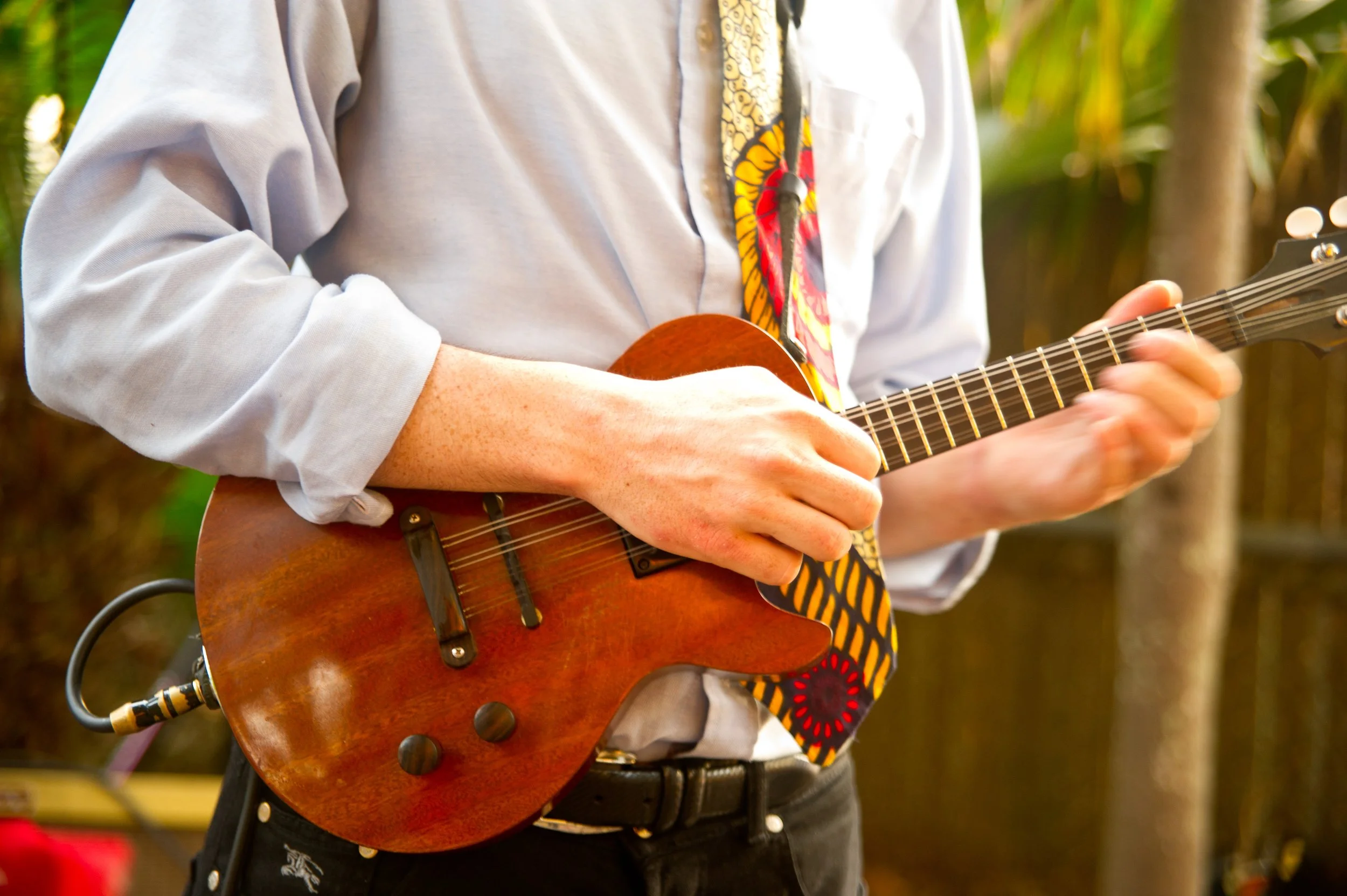 Person playing an acoustic guitar outdoors with a colorful tie and casual gray shirt.