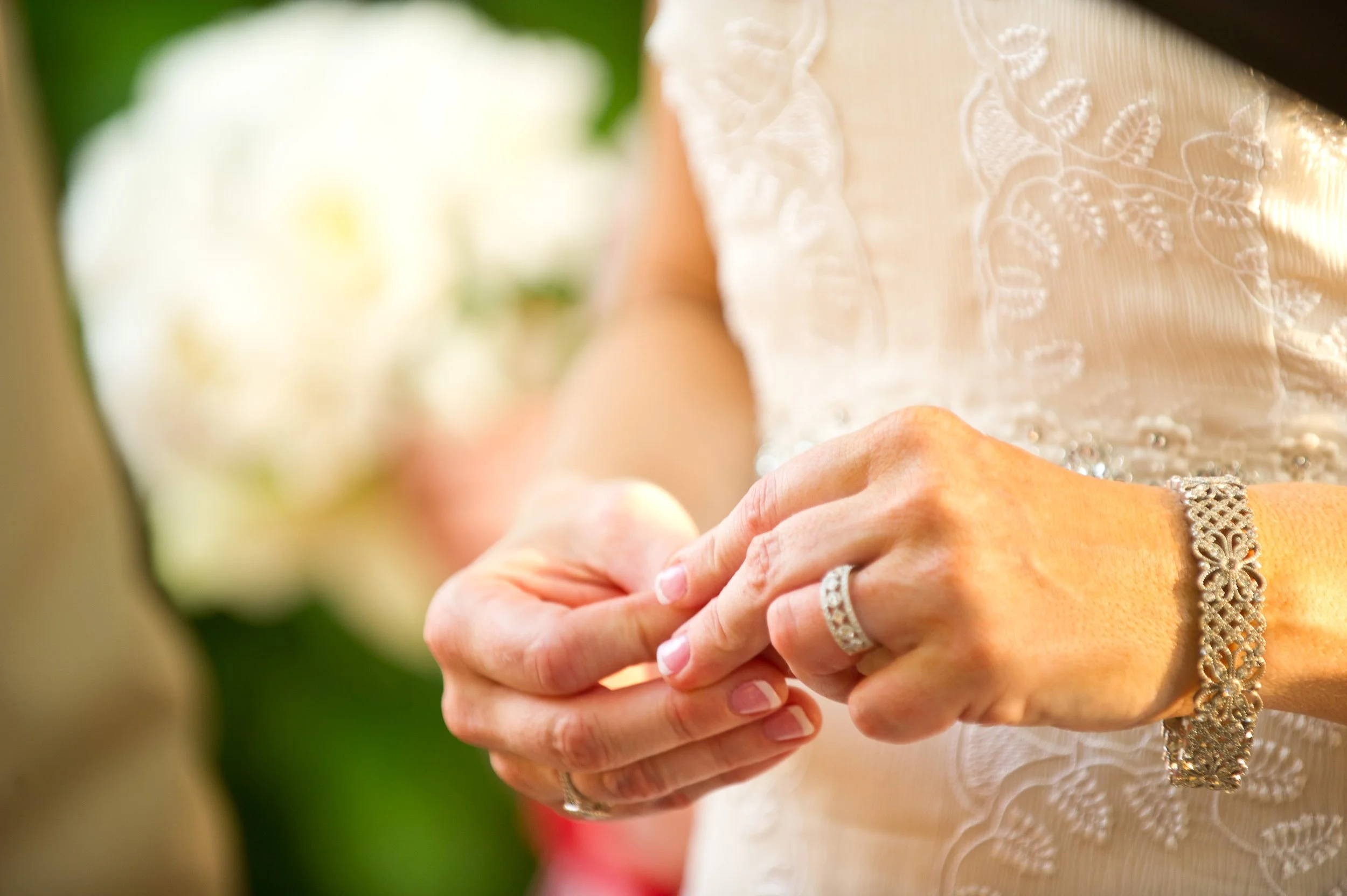 Close-up of a woman's hands holding another person's hand, wearing jewelry, with a blurred ornate dress and a green background.