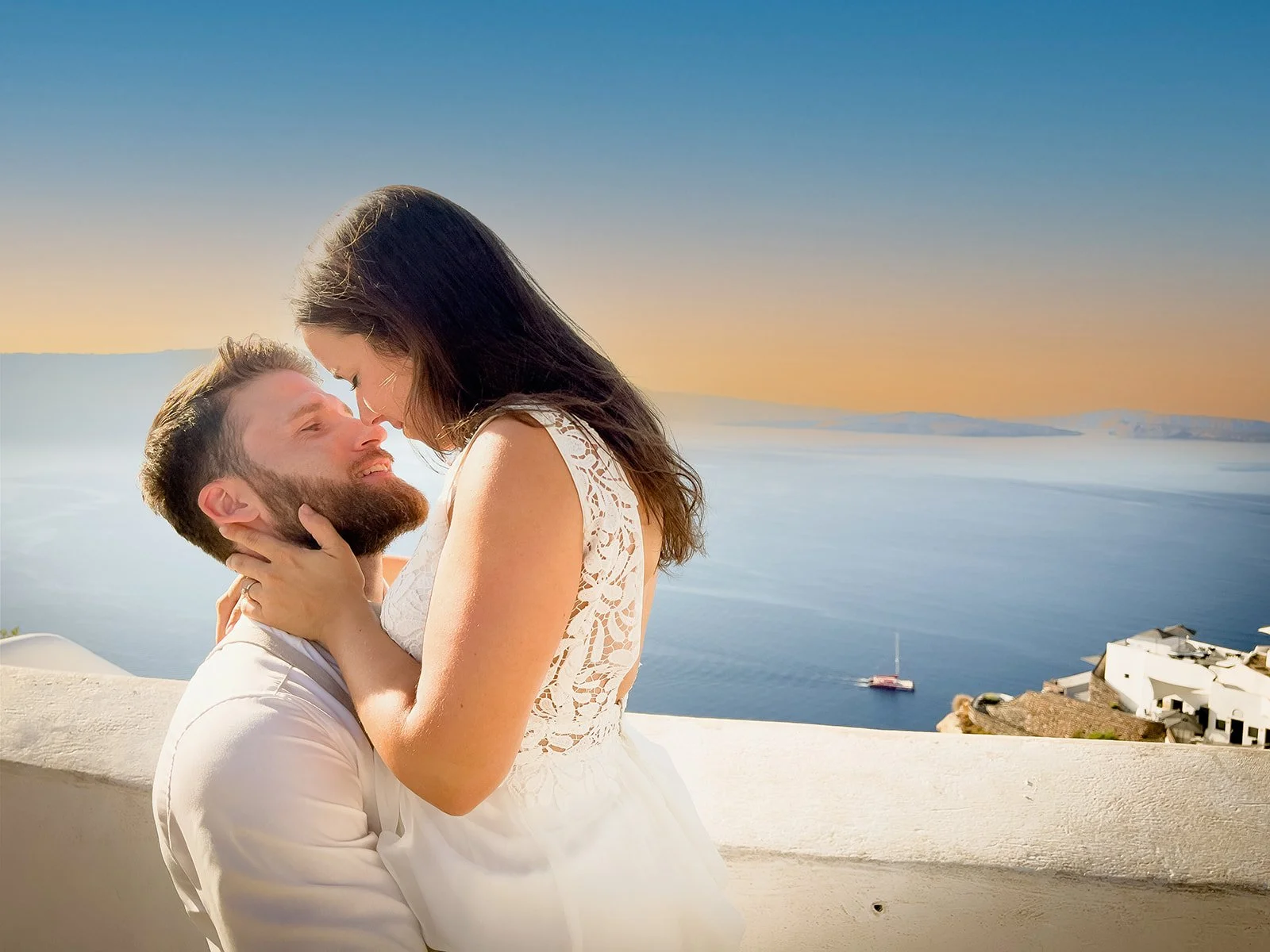 A couple embracing and touching foreheads near a white wall overlooking the ocean at sunset, with water, boats, and white buildings in the background.