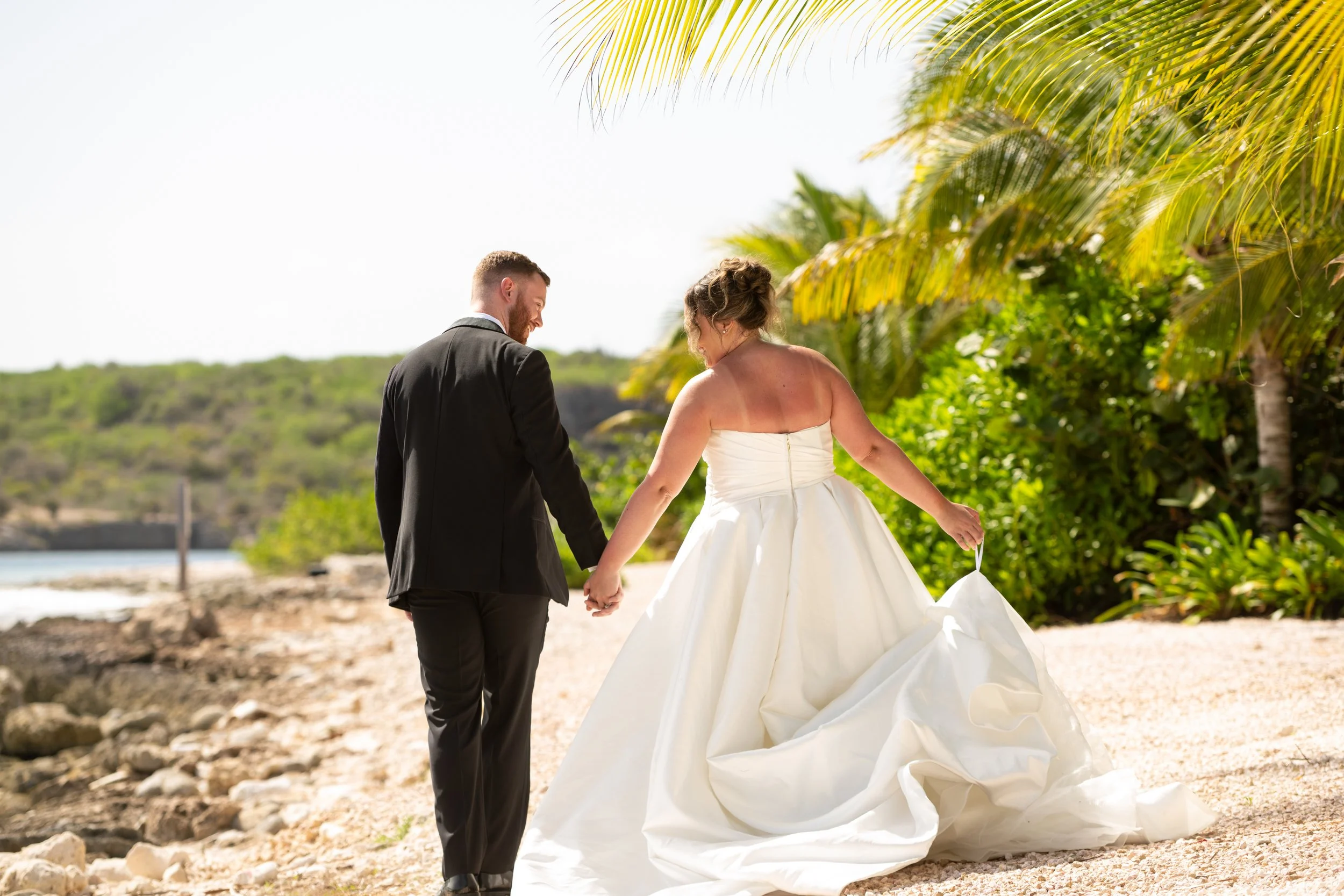 A bride and groom walking hand in hand on a beach with palm trees, during a sunny day.