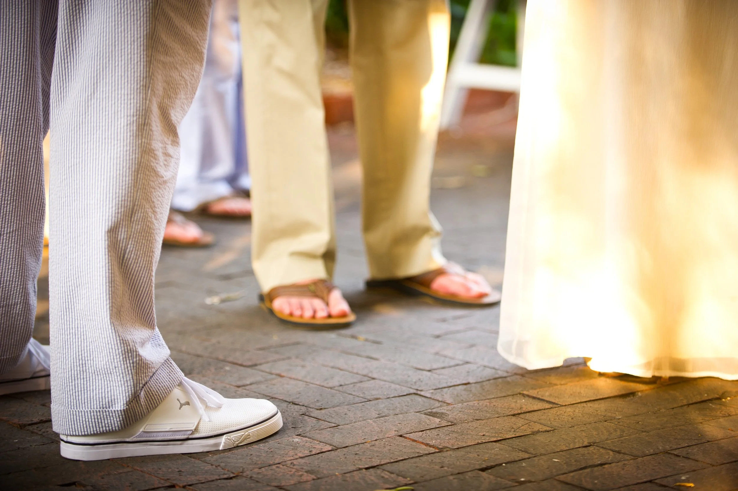 People lined up, standing on a brick sidewalk, wearing sandals and casual clothing, in an outdoor setting with sunlight.