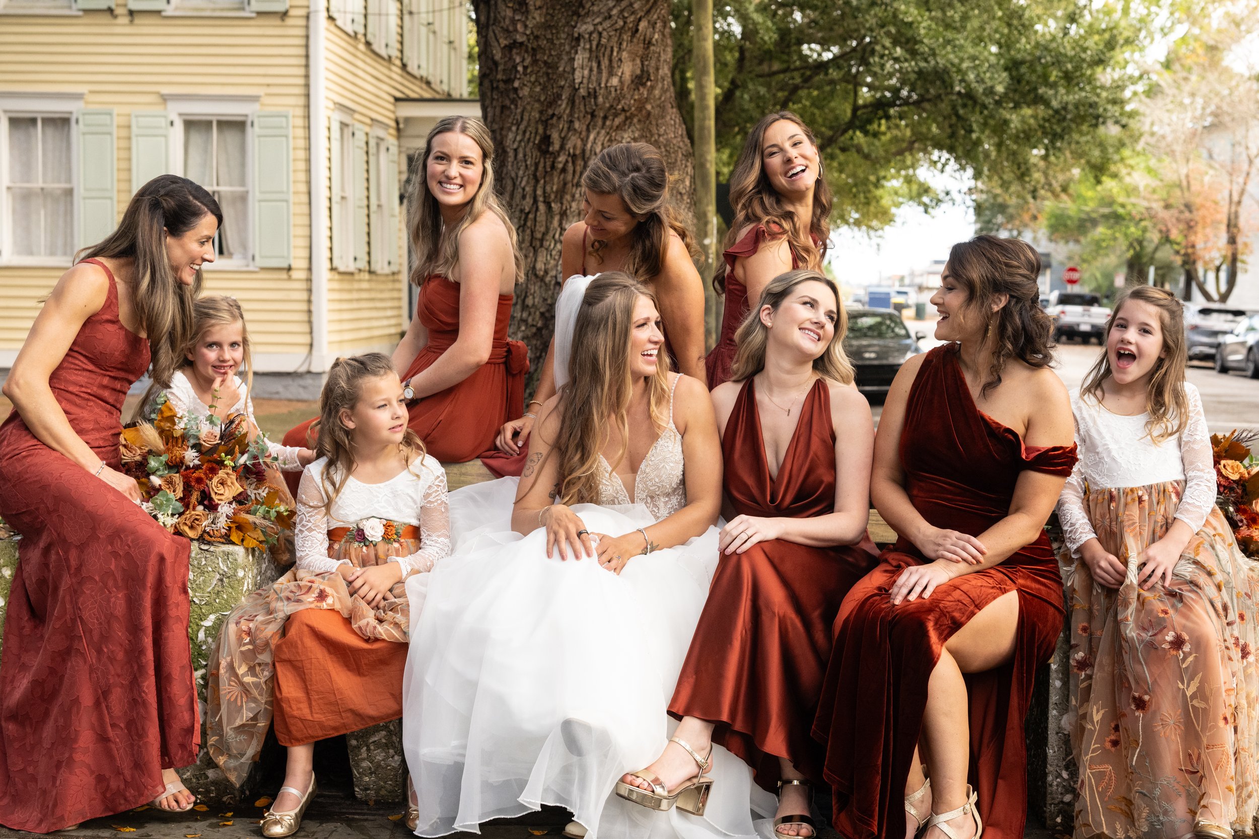 A group of women and children dressed in formal attire, sitting together outdoors near a tree, smiling and laughing during a celebration or event.