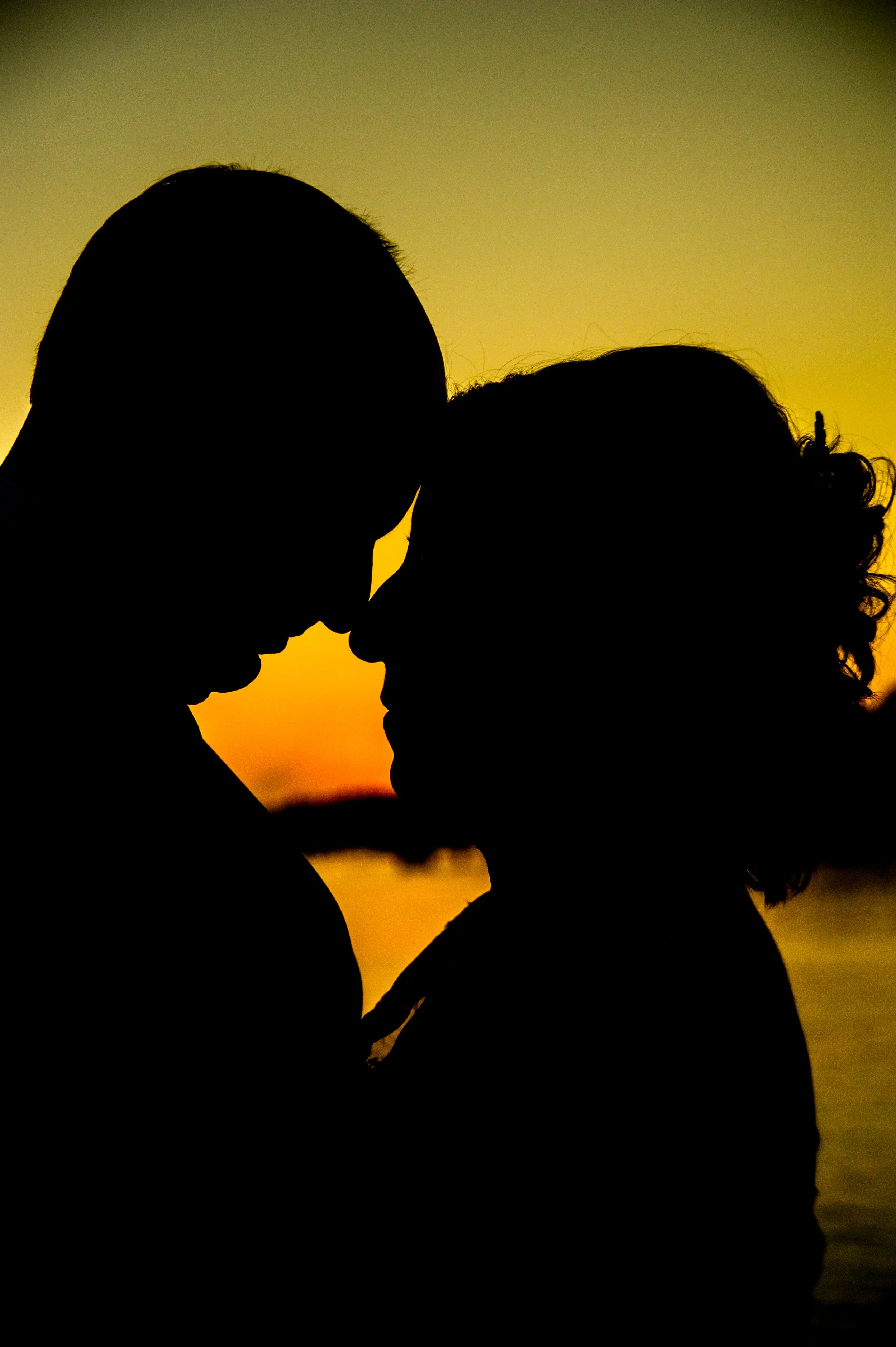 Silhouettes of a man and woman leaning close to each other at sunset, with the orange sky in the background.