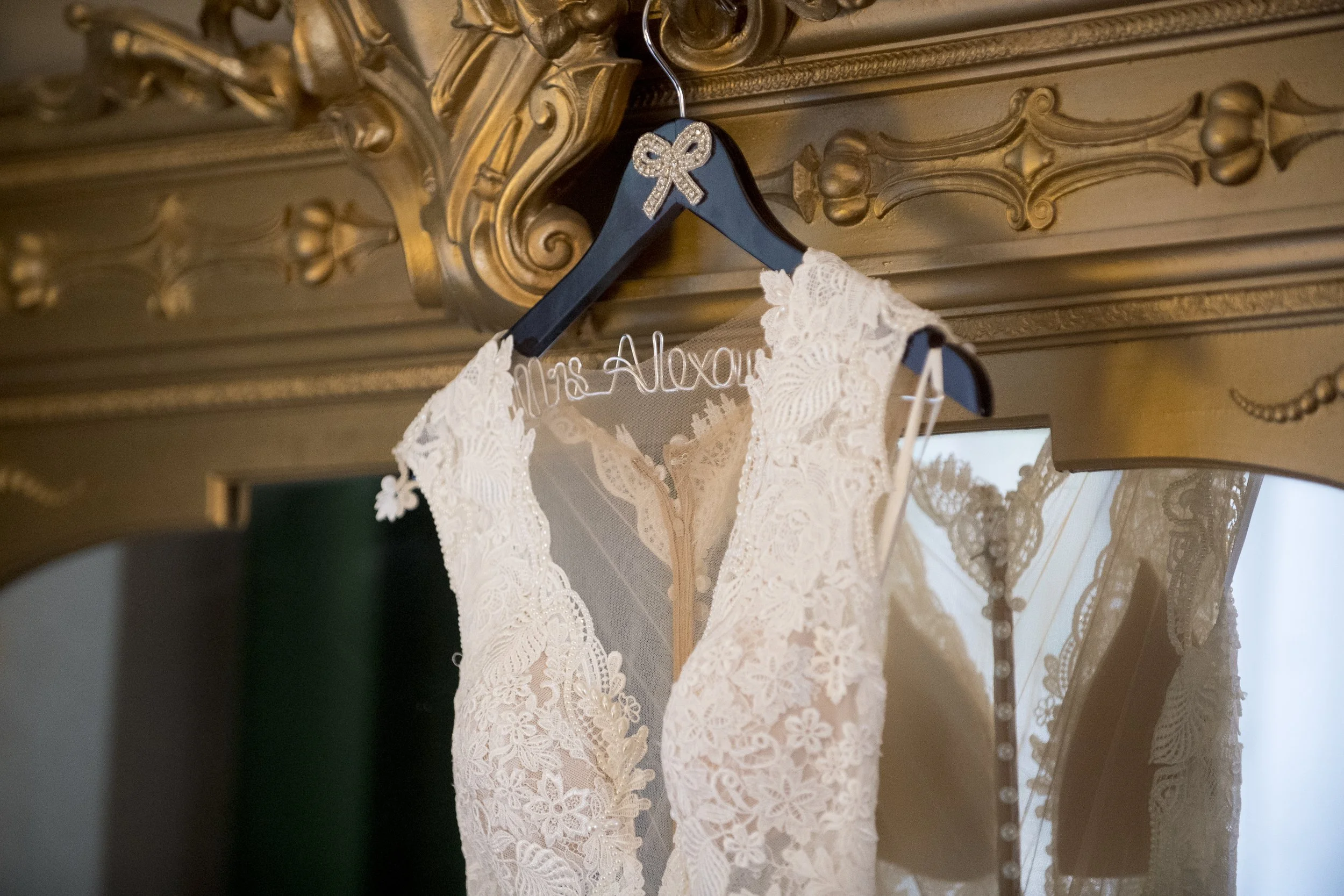 A wedding dress hangs on a decorative black hanger with a rhinestone bow, inside a wooden wardrobe with elaborate gold carvings. The dress is made of lace with floral patterns and has a high neckline, displayed against a mirror and a window.