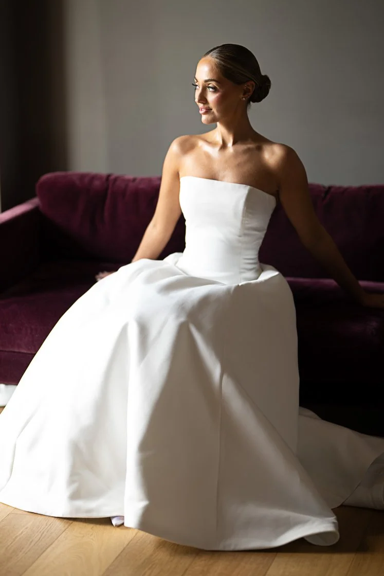 A woman in a strapless white wedding gown sitting on a purple sofa, looking to her right.
