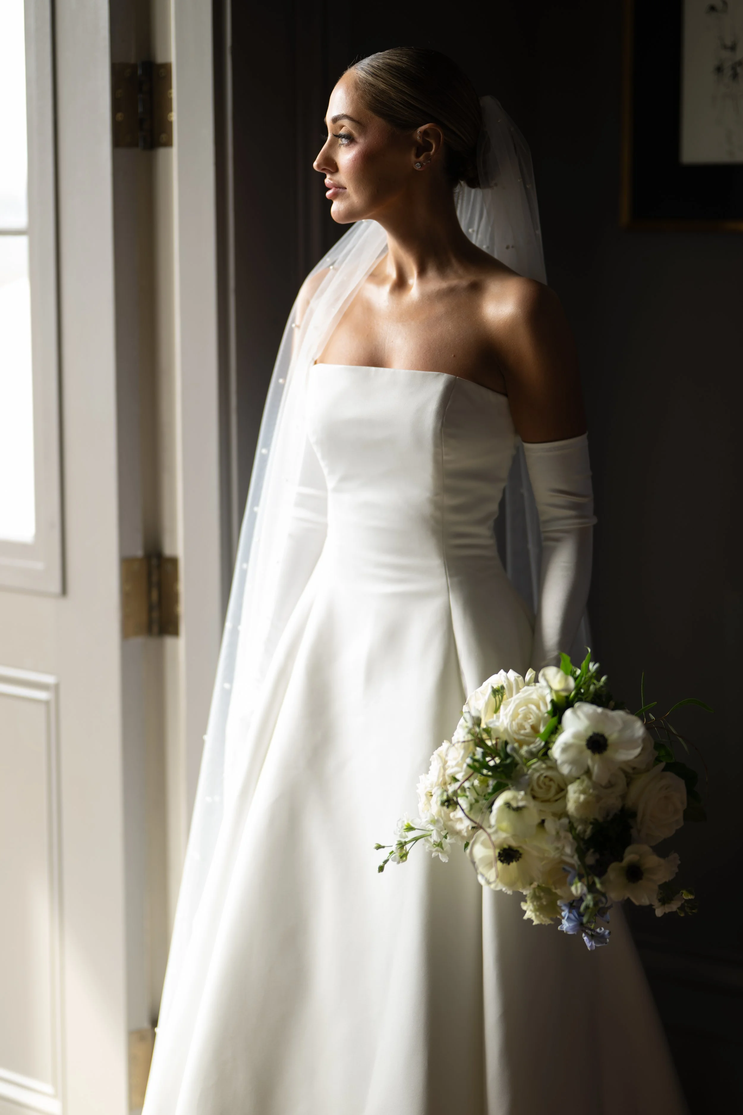 A bride looking out a window, wearing a strapless white wedding gown, a veil, and earrings, holding a bouquet of white and dark flowers.