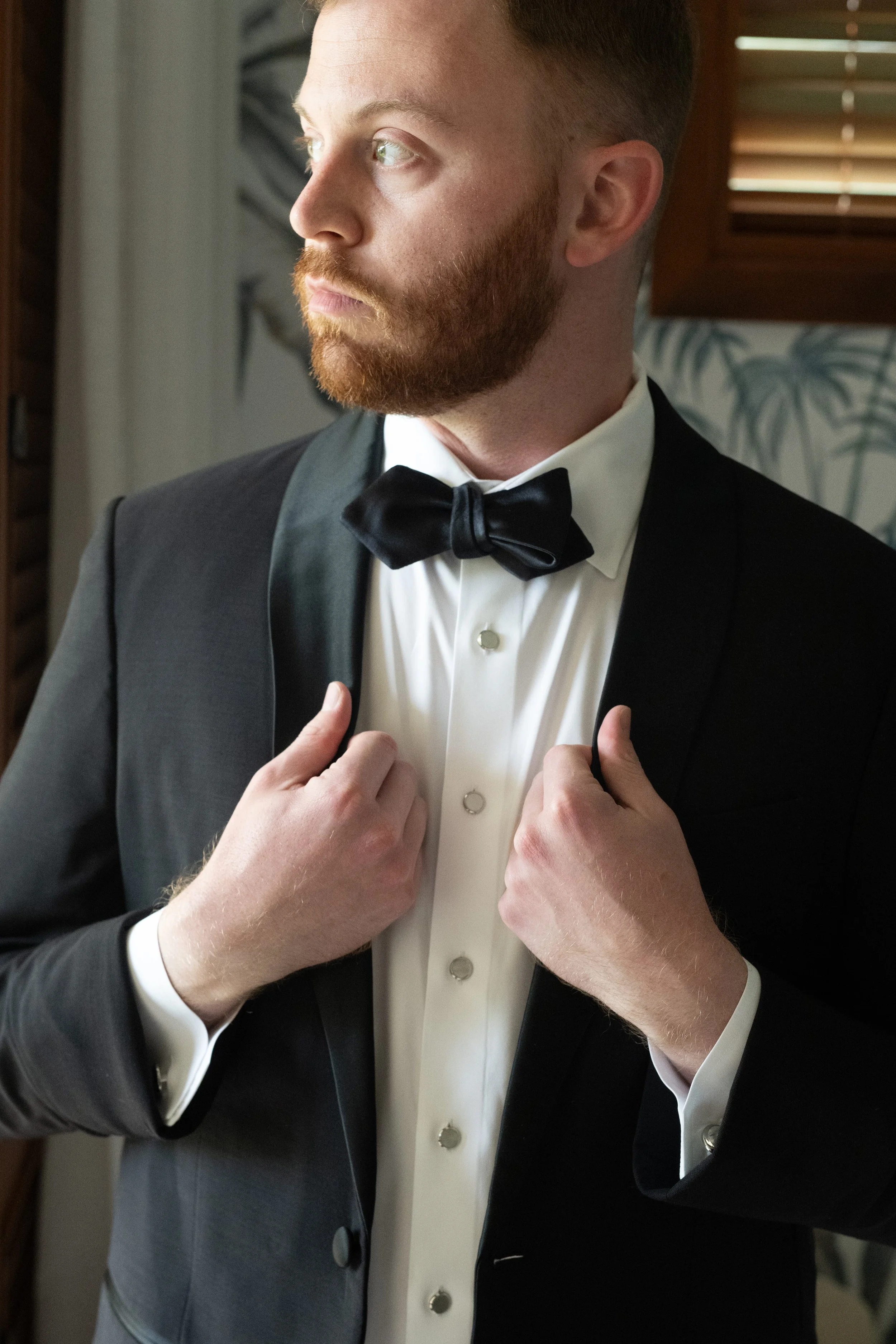 A man in a tuxedo adjusting his tuxedo jacket near a window with blinds.